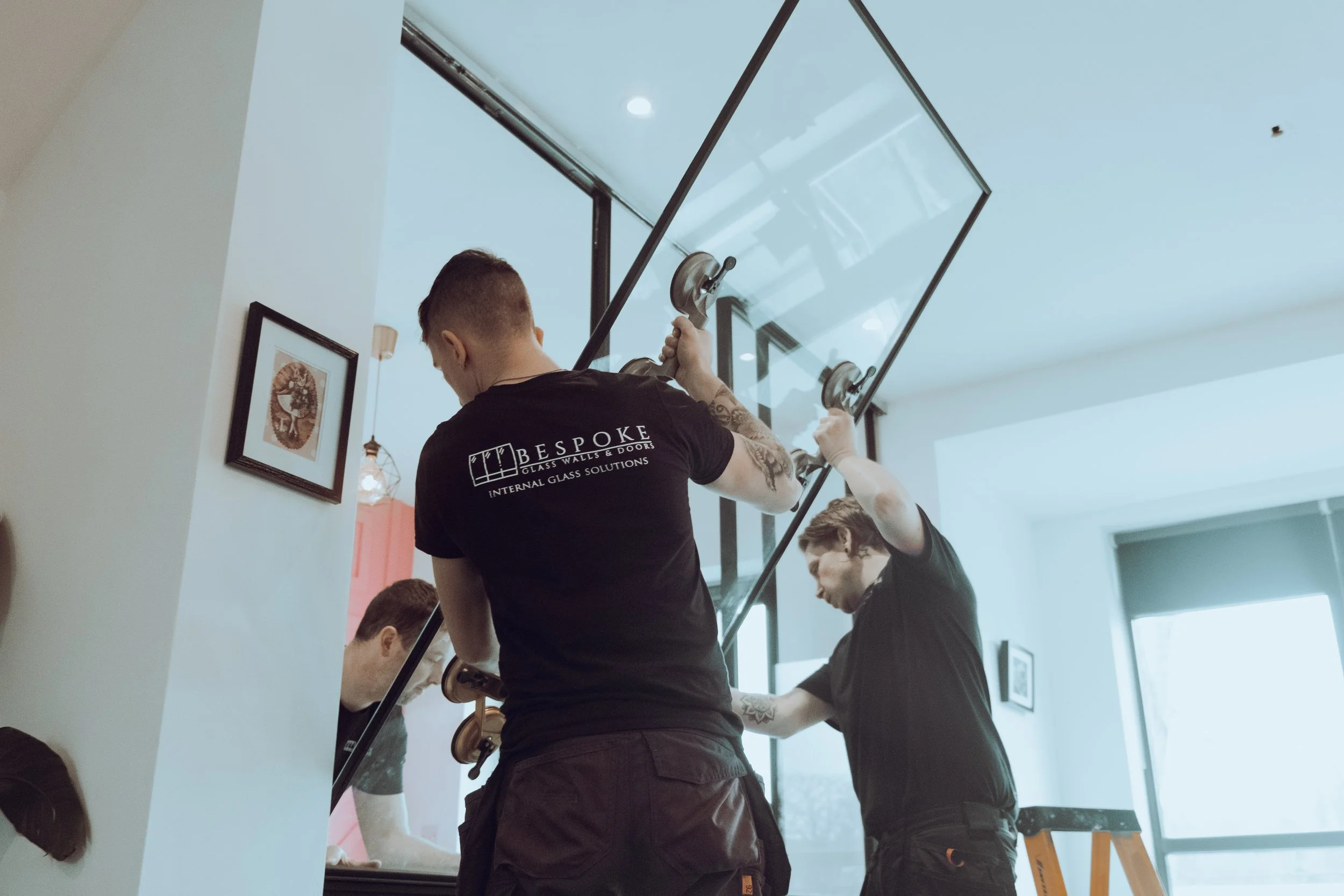 Two fitters in branded tee shirts lifting a large glazed panel with vacuum suction cups into a black steel frame inside a house.