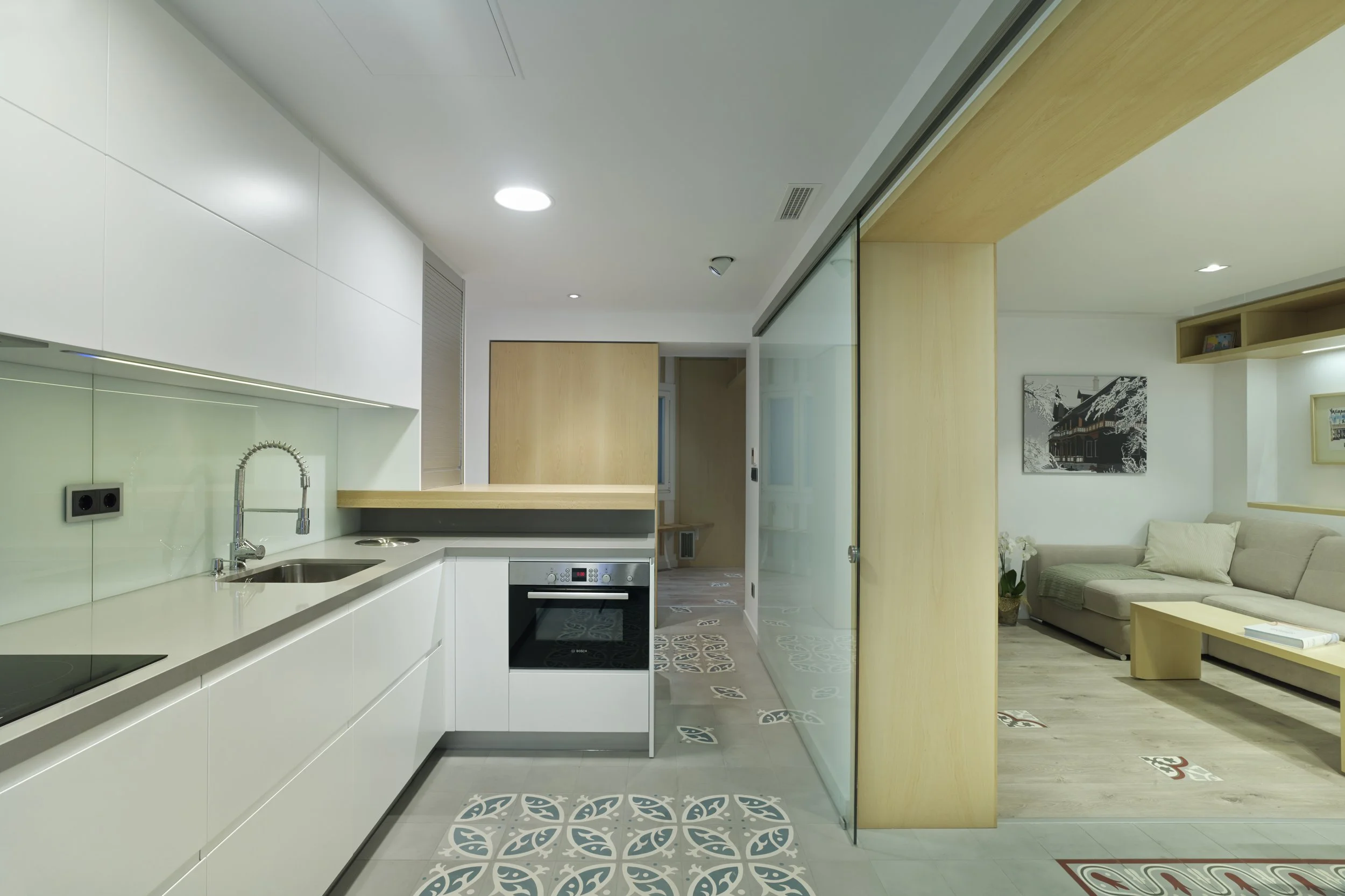 Modern kitchen with white cabinets, a black oven, and a sink, adjacent to a living room with a beige sofa and framed artwork, separated by a glass sliding door.