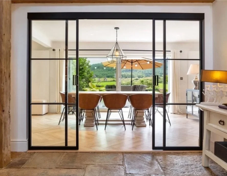 A stone floored hall looking through black steel framed double sliding doors with fixed sidelights into a bright dining room with a garden beyond.