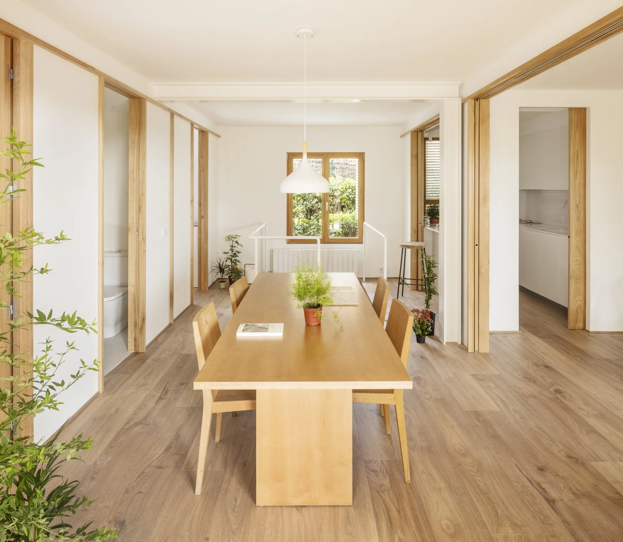 Bright dining room with wooden table and chairs, plants, large window, and partial view of bathroom and kitchen.