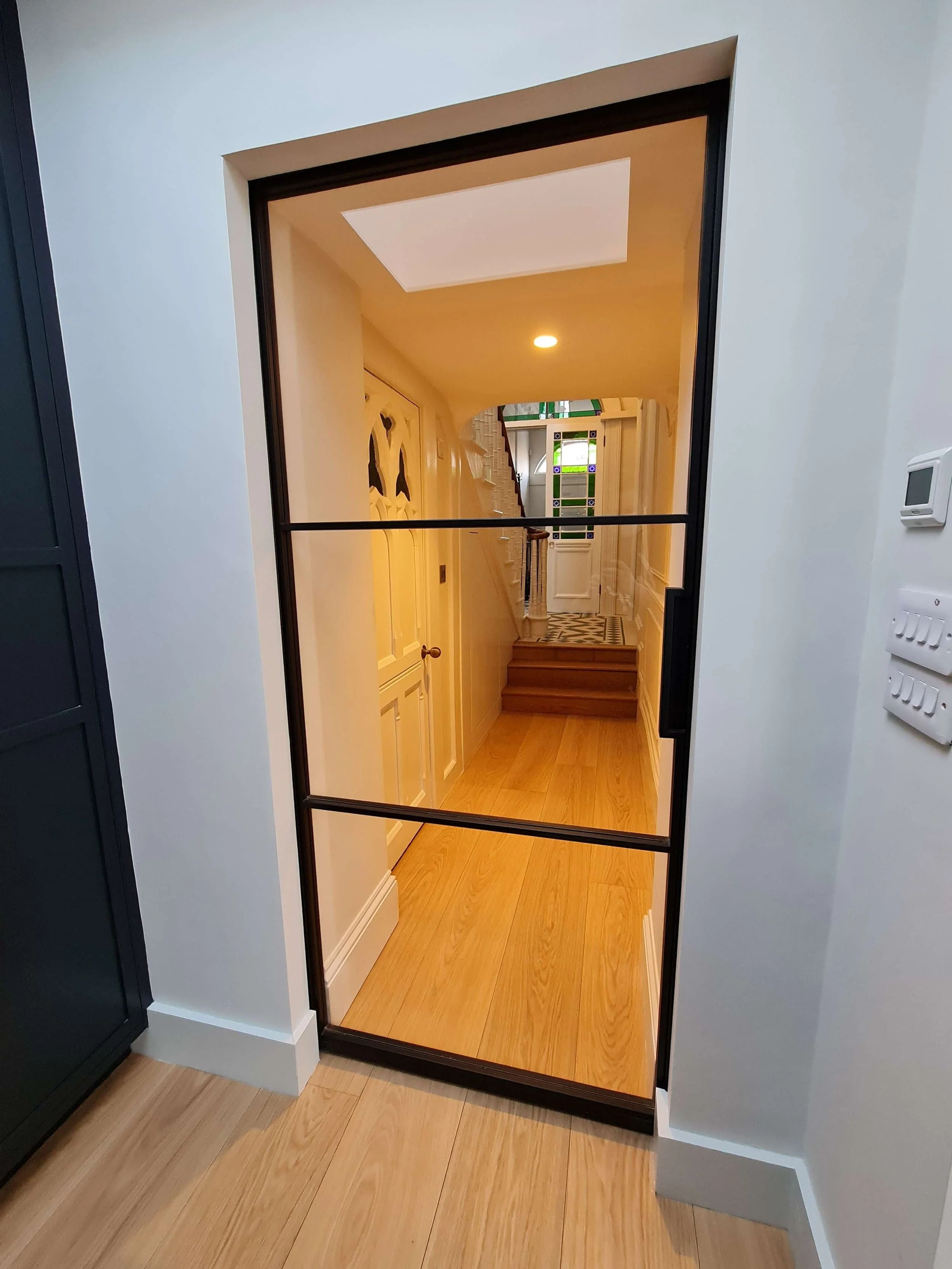 View through a glass door into a hallway with wooden flooring, white walls, and a staircase leading upstairs.