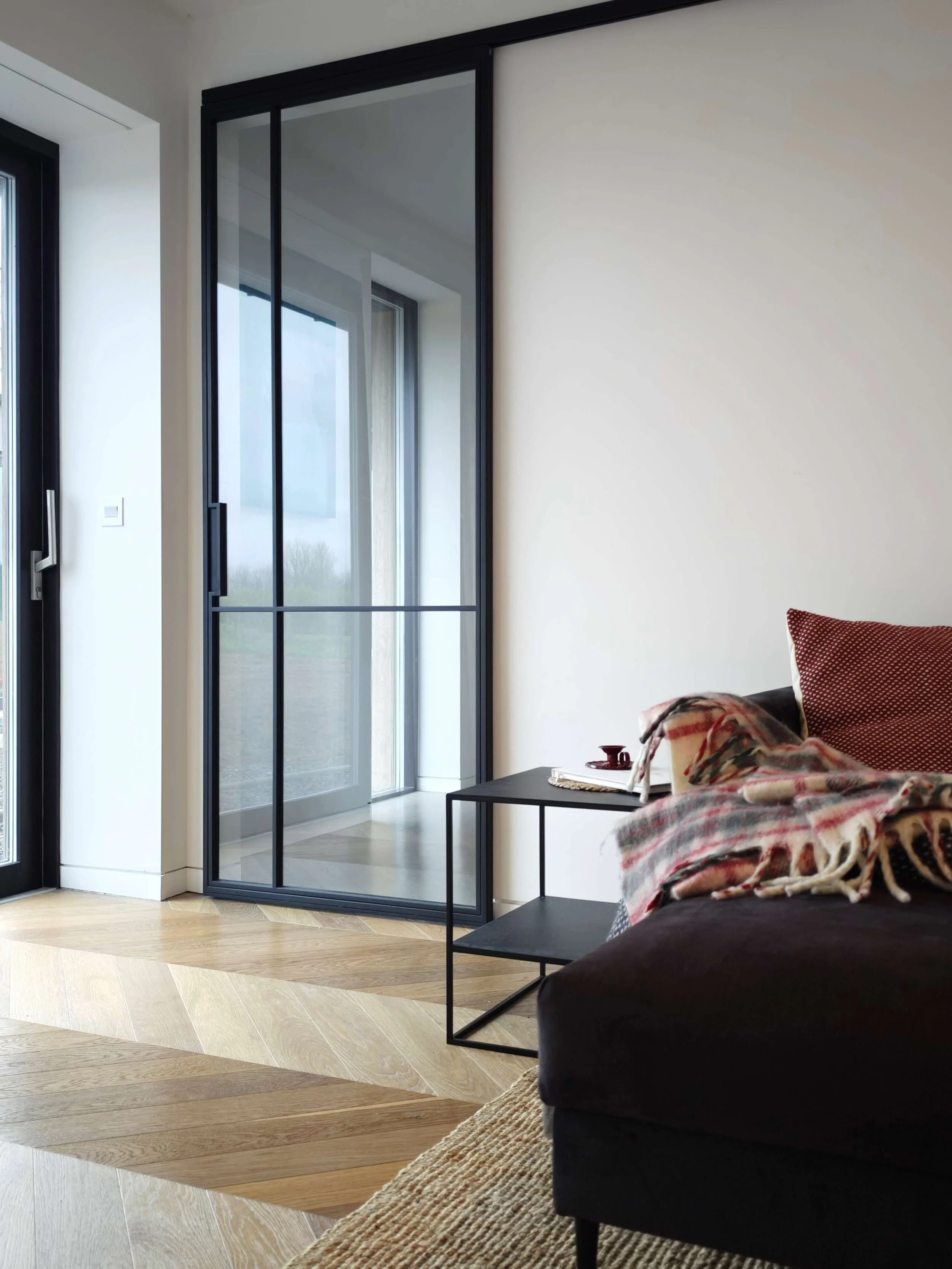Minimalist living room with a dark sofa, patterned throw, black side table, and large glass sliding door letting in natural light.