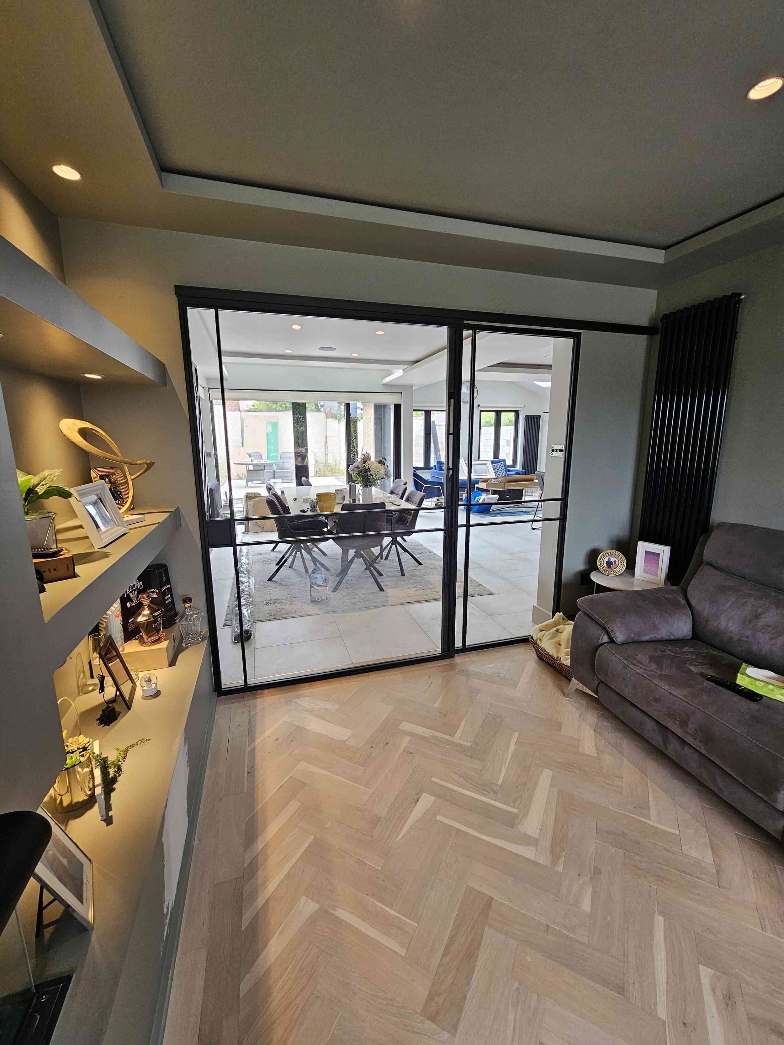 Modern living room with glass doors leading to a bright dining area, shelves on the left, and a brown recliner on the right.