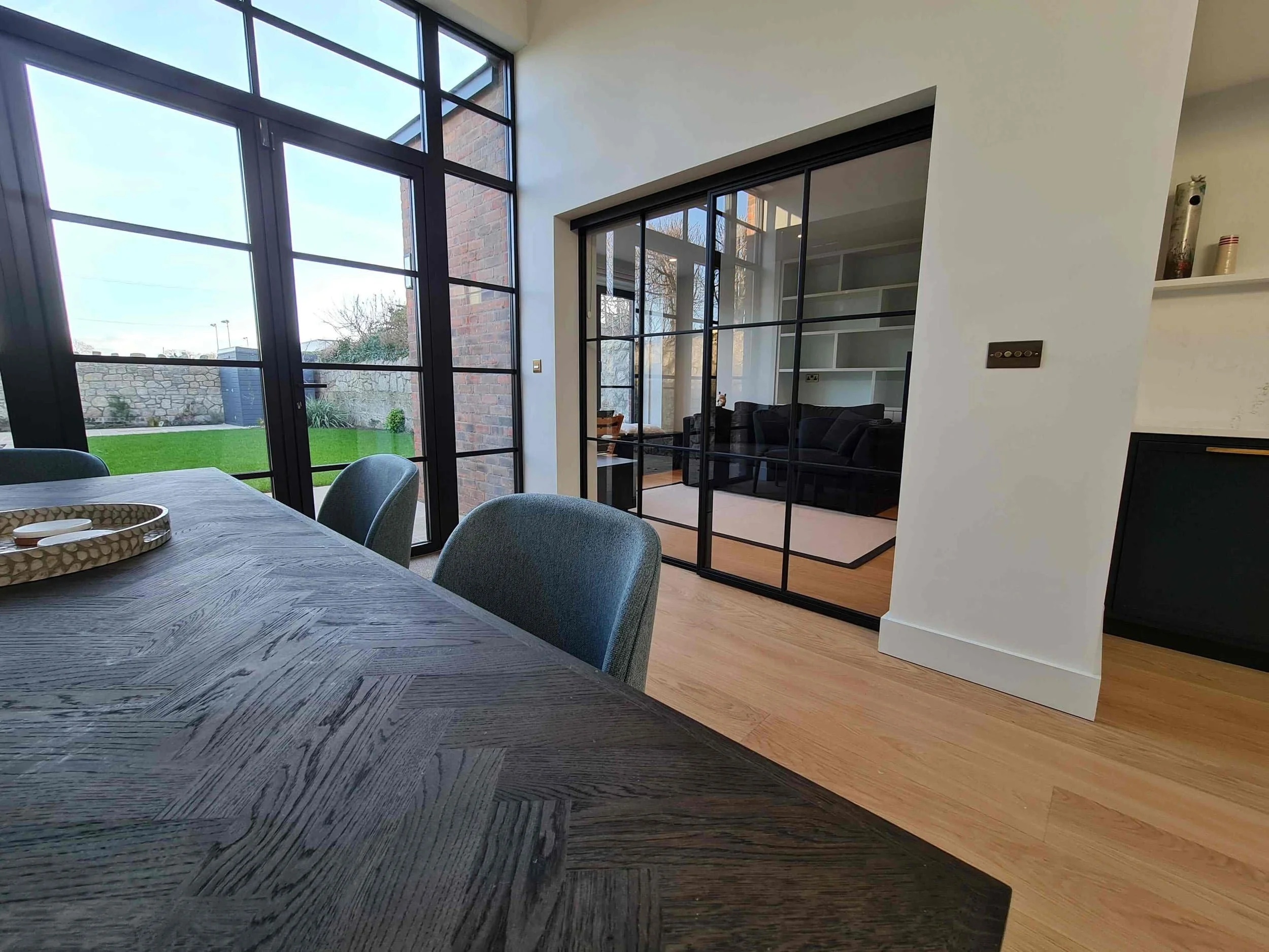 Modern dining area with dark wood table, glass doors, and view into living room with shelves and black sofa; large windows.