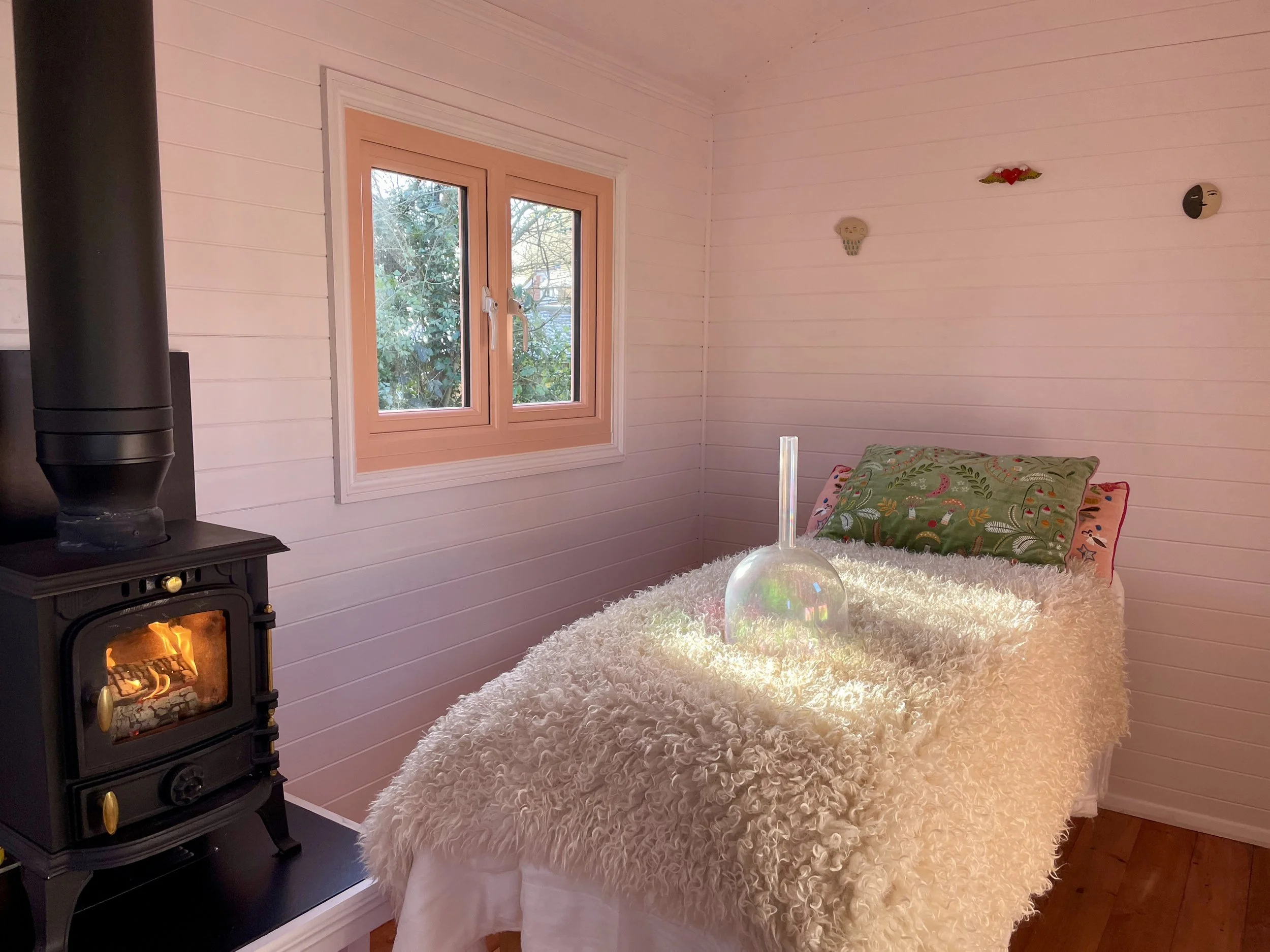 A cozy therapy room with a bed covered in a fluffy white blanket, decorative pillows, a glass vase with a straw, a wood stove with a visible fire, and a window with a view of trees outside.