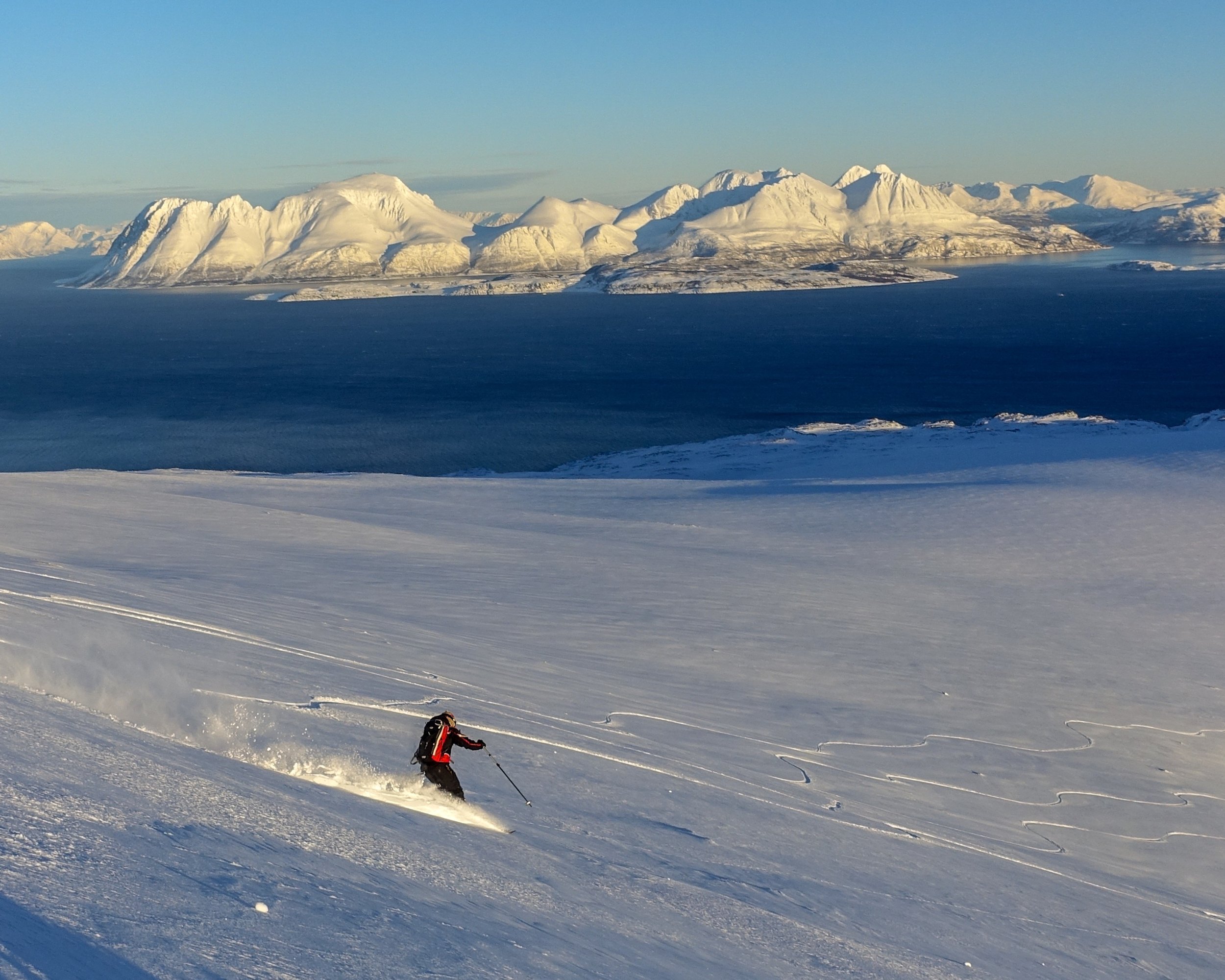 Skiløper nedover snødekt skråning med fjell og hav i bakgrunnen