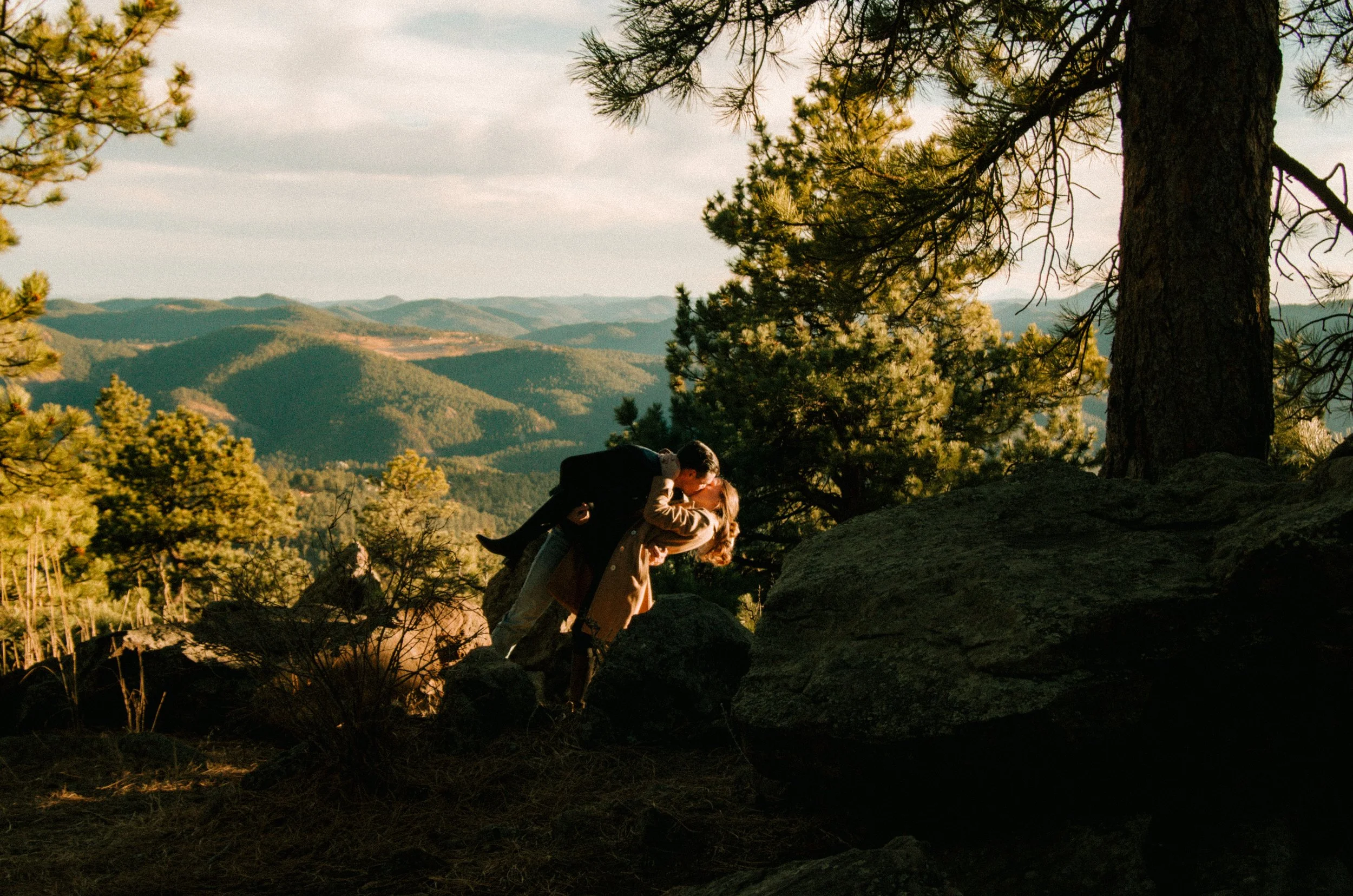 A couple kissing on a rocky ledge in a forested mountainous area during sunset.