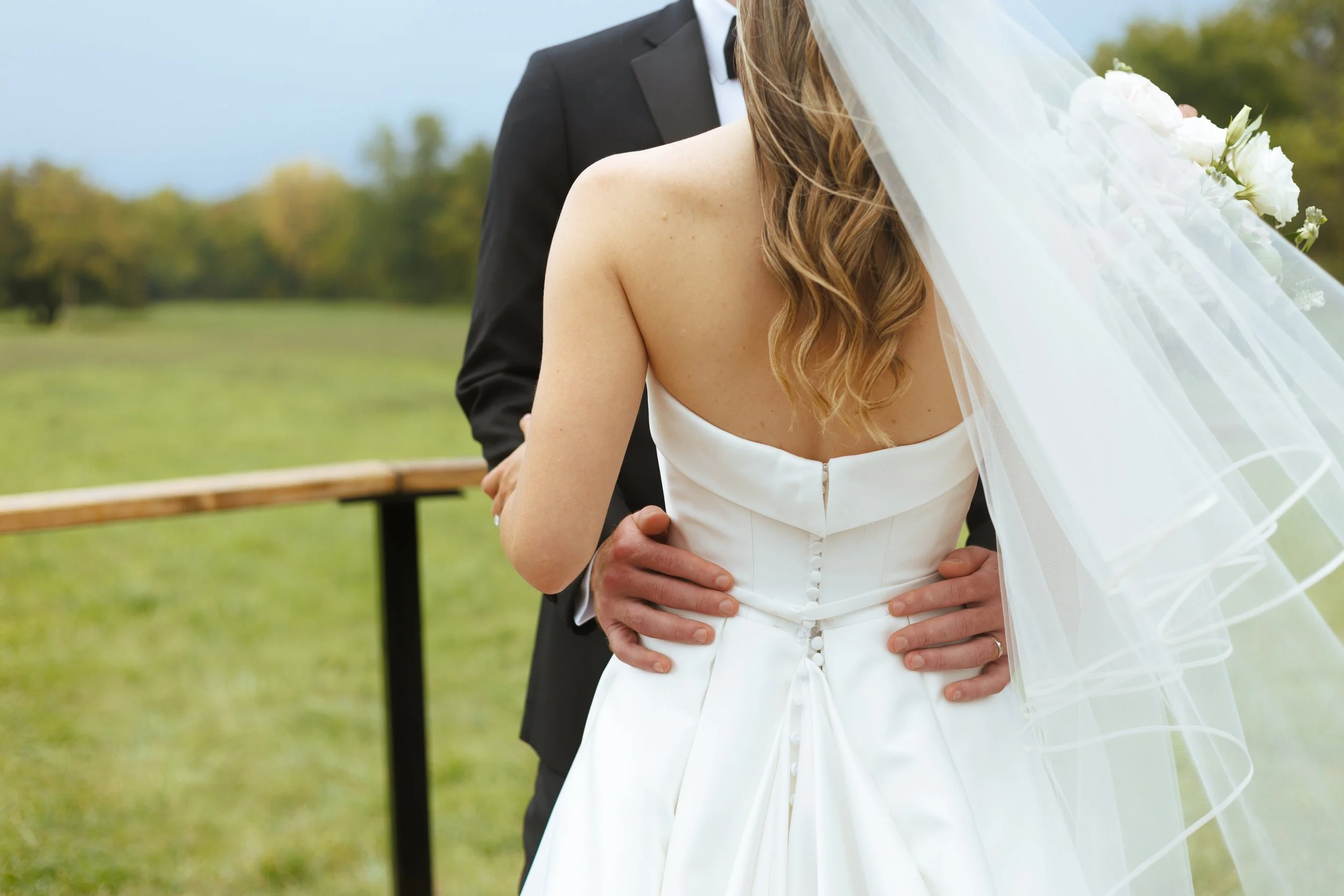 Bride and groom holding each other outdoors during wedding ceremony, with the bride in a white strapless gown and veil, and the groom in a black suit.