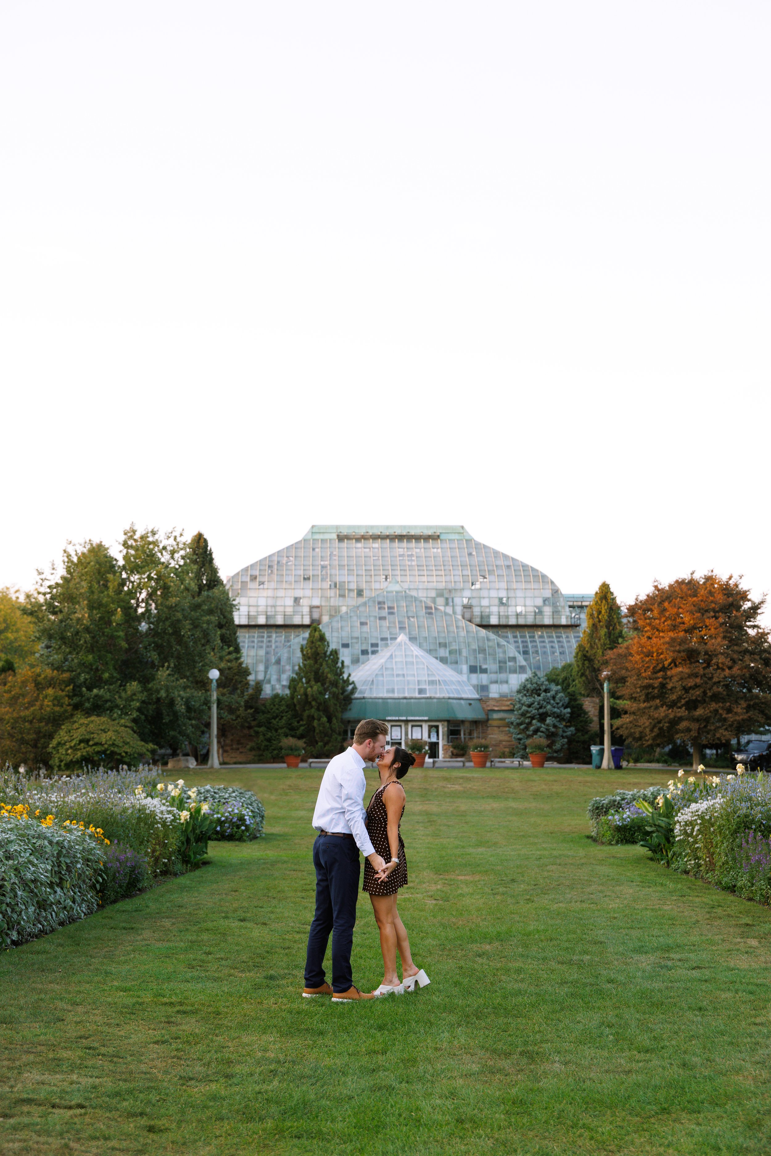 A couple holding hands and about to kiss in a garden with a large greenhouse in the background.