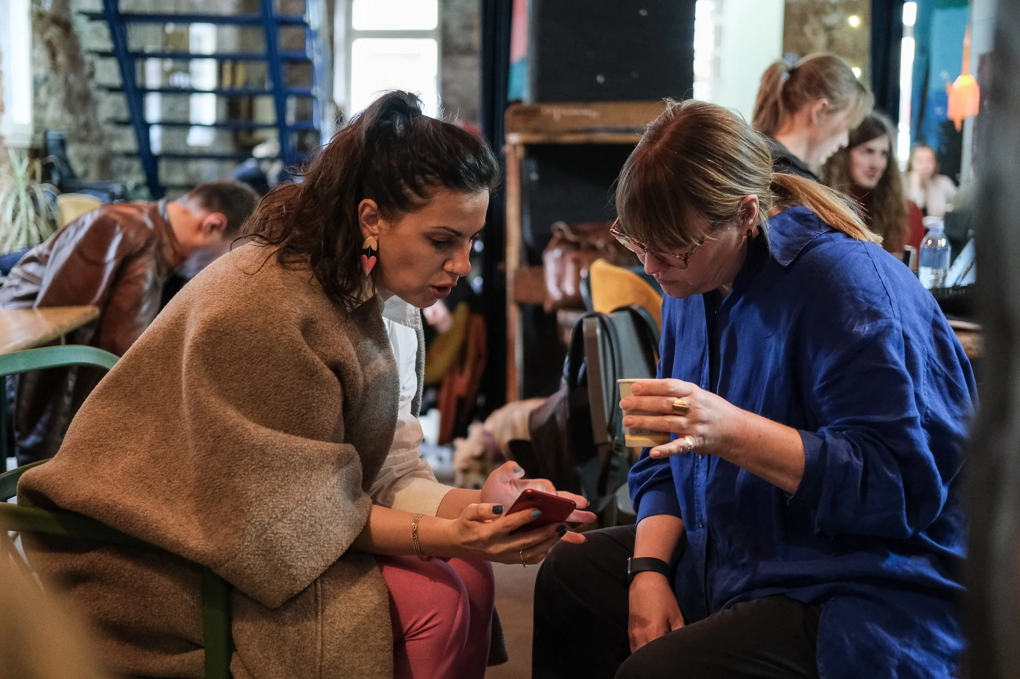 Two women seated closely together in conversation inside a rustic, industrial-style space with exposed brick walls and colorful artwork. One woman, with brown curly hair, is wearing a beige coat, and the other, with straight hair and glasses, is wearing a blue shirt. They appear to be engaged in a serious discussion.