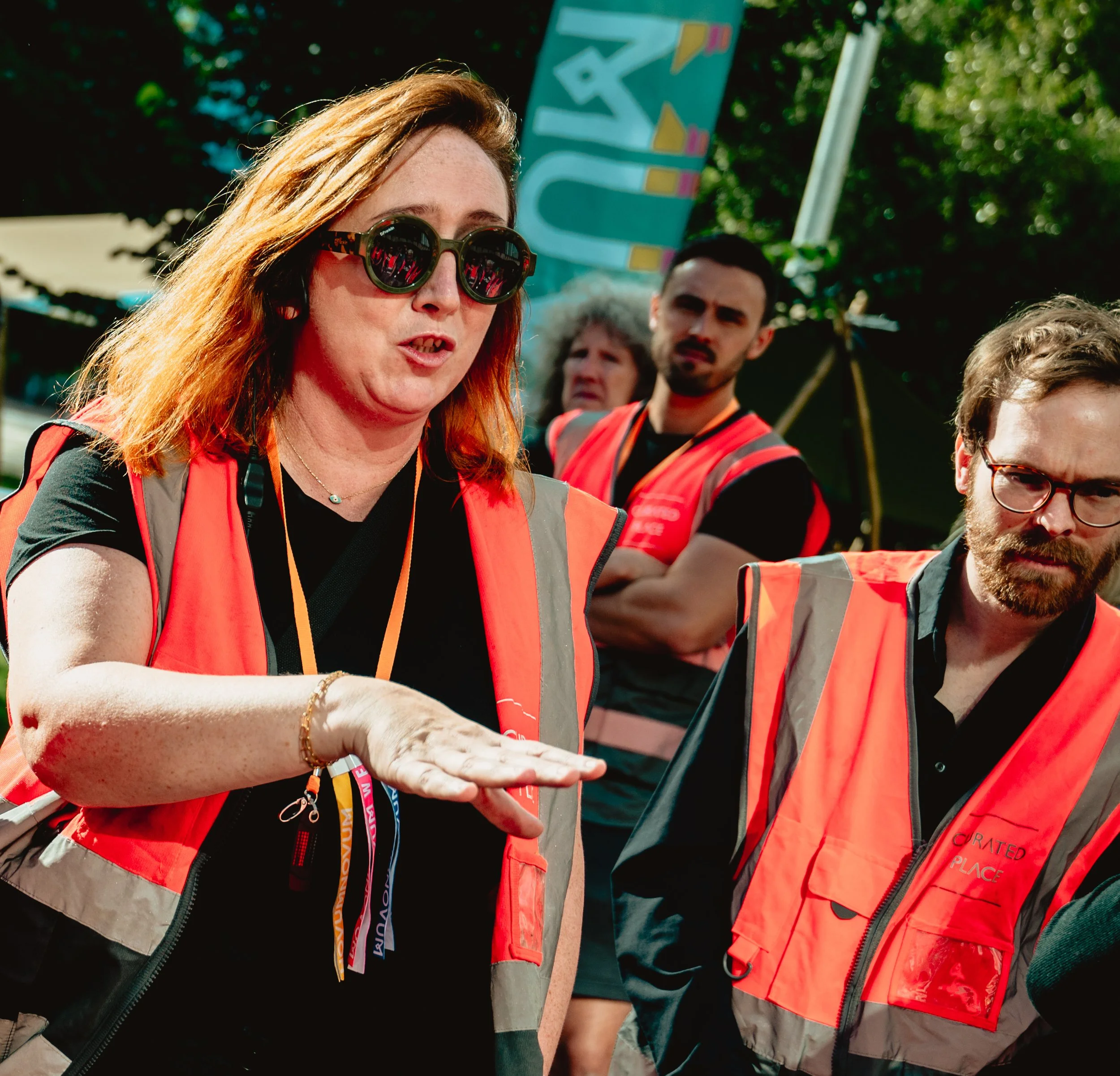 Three people in hi-vis vests at an outdoor event in the sun. One person talking to the other, gesturing