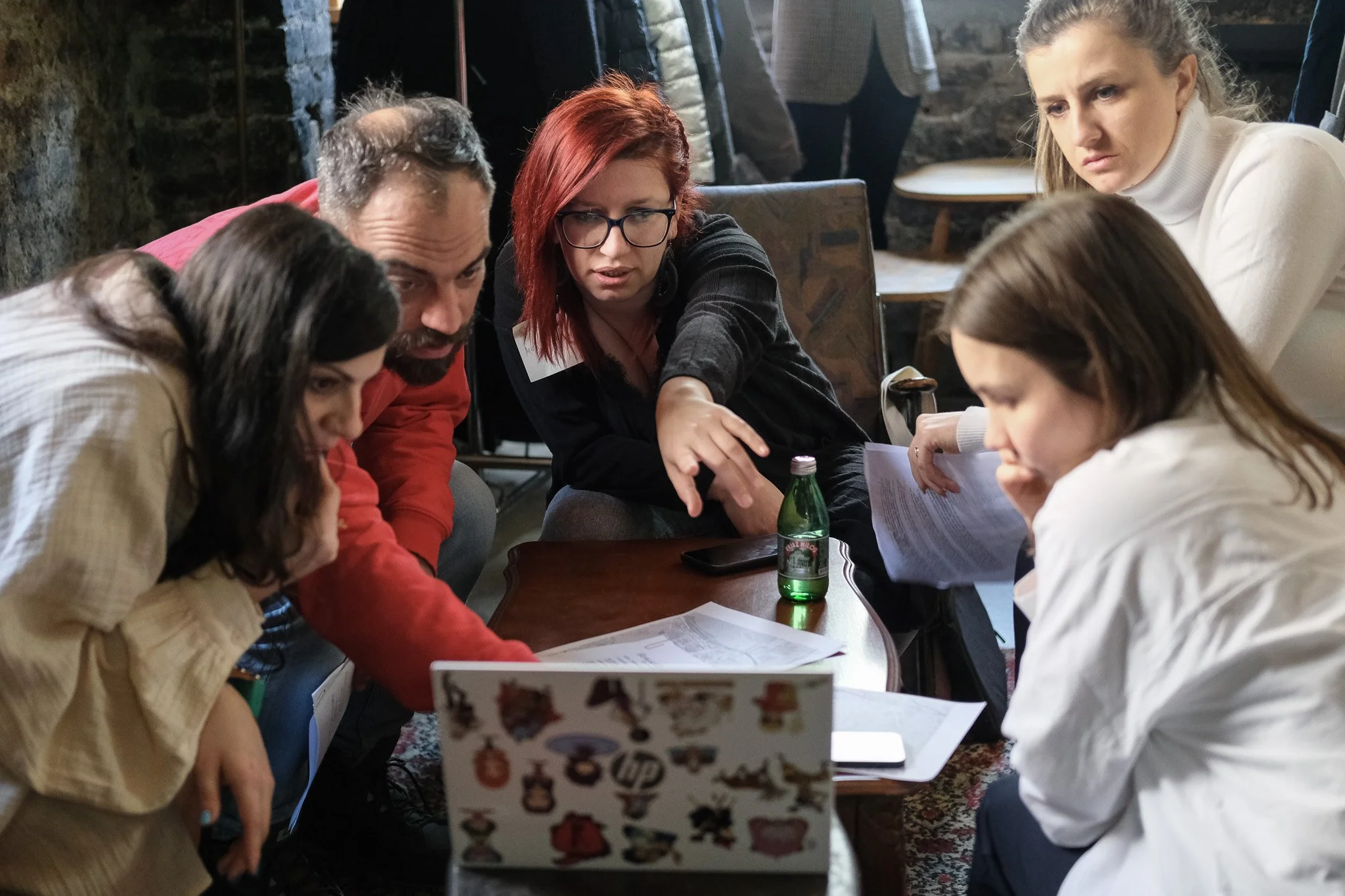 A group of five people gathered around a table, looking at papers and a laptop, in a cozy room with stone walls. A woman with blonde hair and a white turtleneck appears to be leading the discussion.