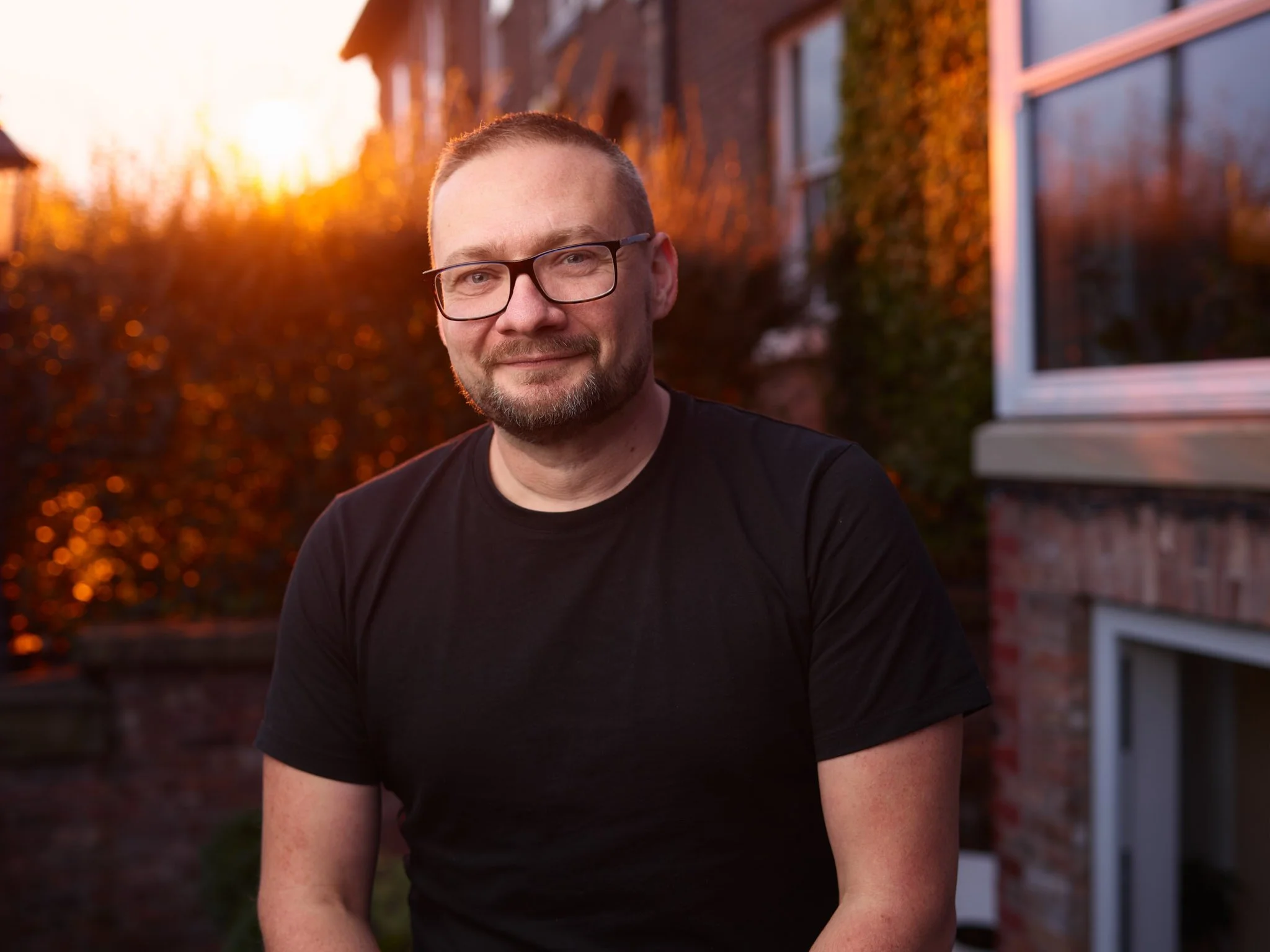 A man with glasses and a beard smiling outdoors during sunset