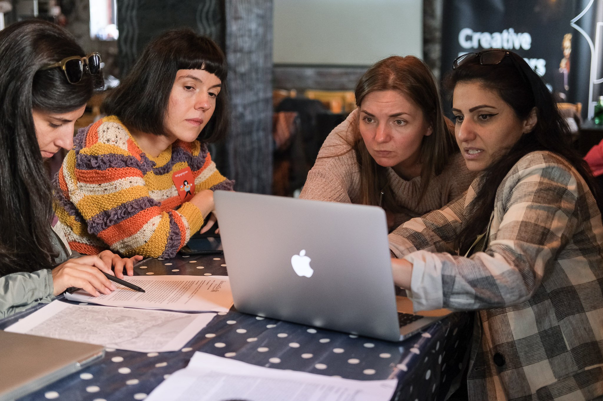 Four women sit at a table looking at a laptop screen, engaged in a discussion. The table has papers, a pen, and a silver laptop. They are in a modern indoor space with a dark background and a sign that reads 'Creative'.