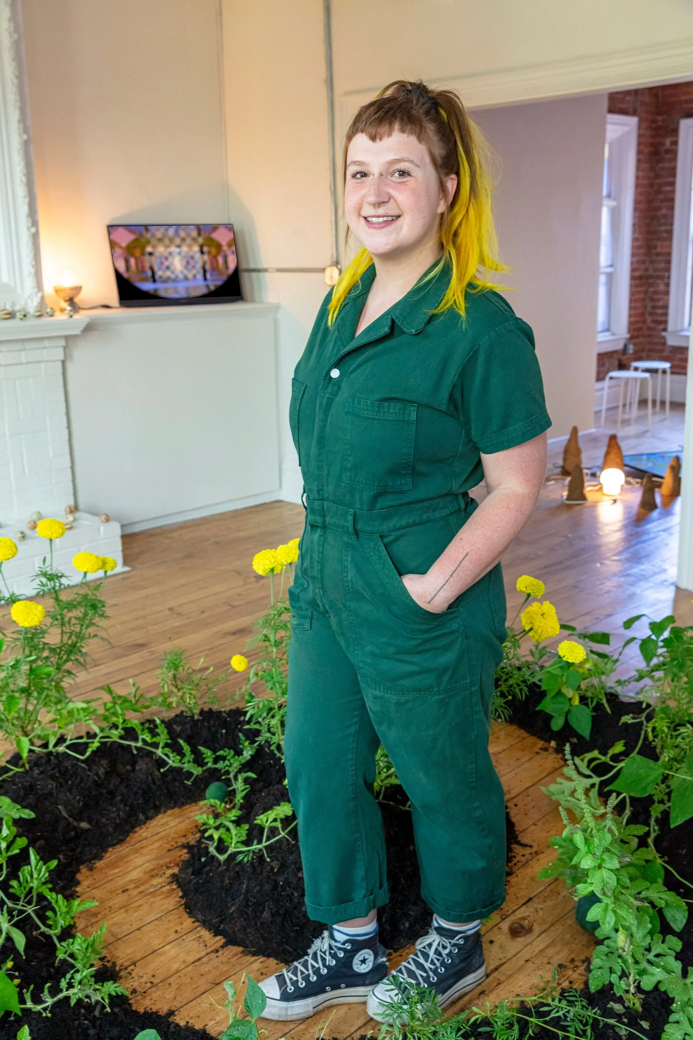 Lucia Riffel stands and smiles in front of their art. They are wearing a green jumpsuit and have their hands in their pockets. They are inside and it is daytime.