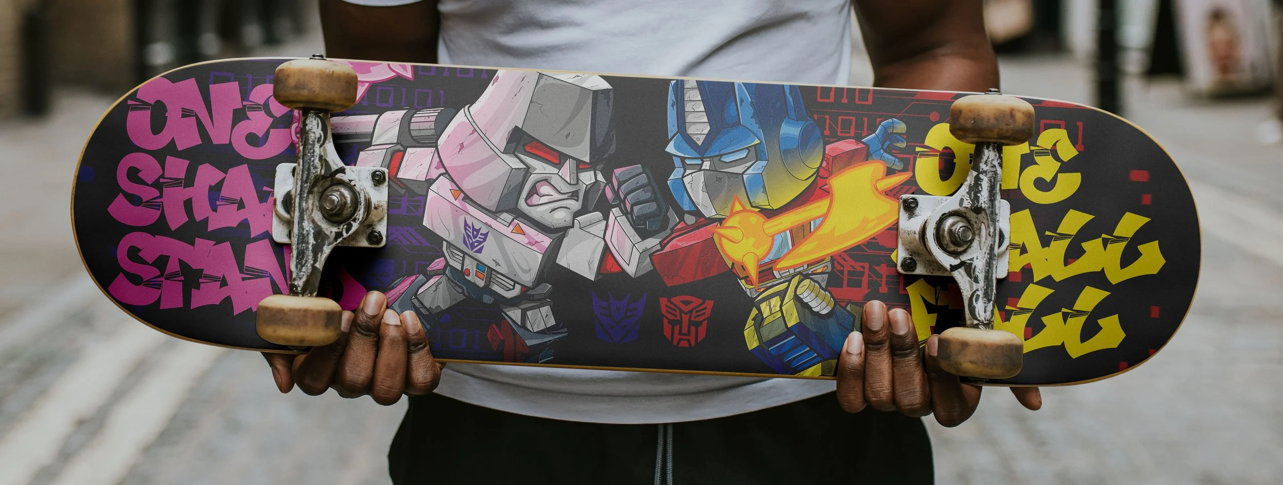 Skateboard featuring Transformers vector graphic artwork of Optimus Prime battling Megatron in a Chibi art style.