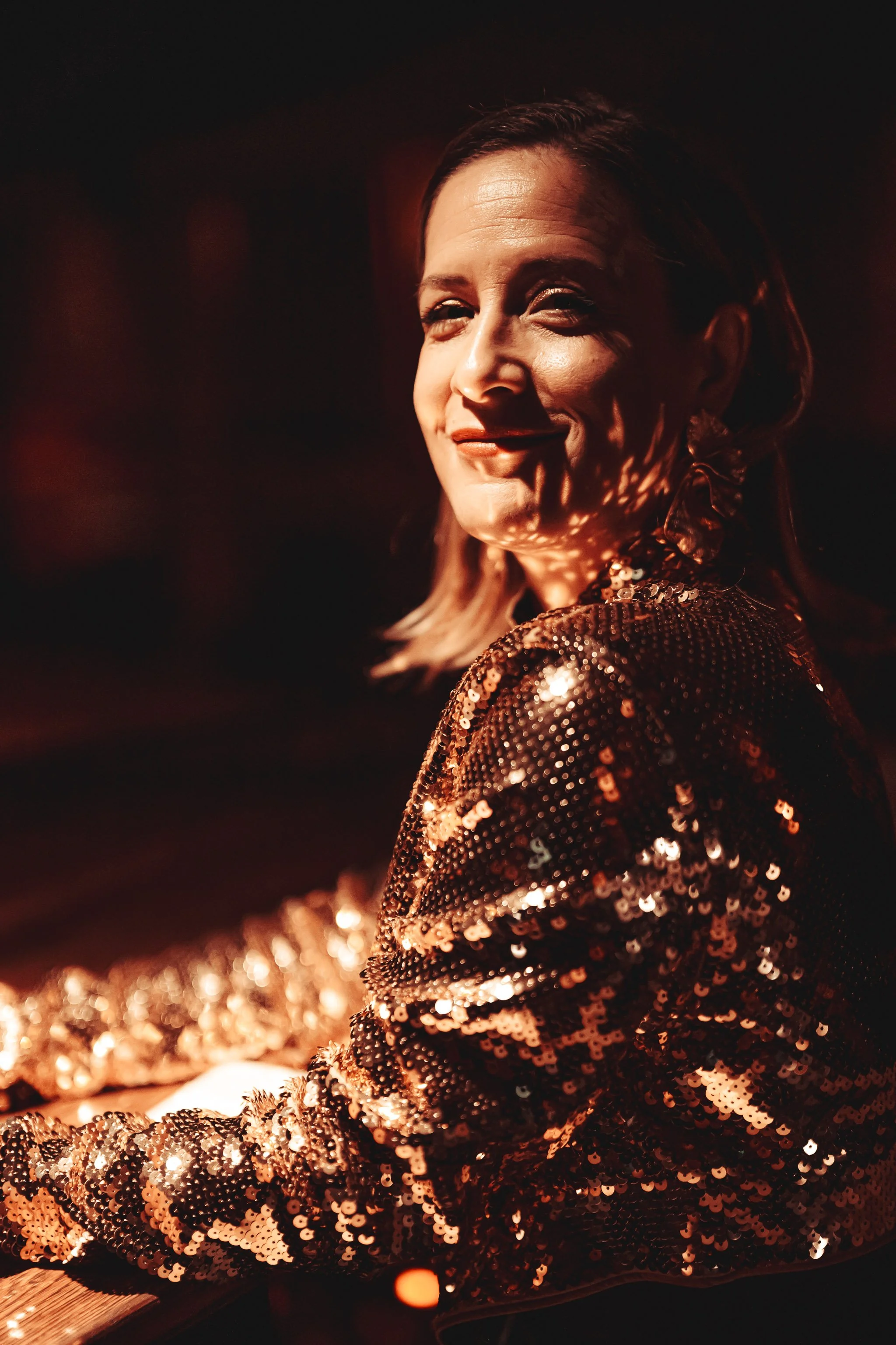 A woman with shoulder-length hair wearing a shiny, sequined, gold-colored jacket, smiling in a dimly lit environment.