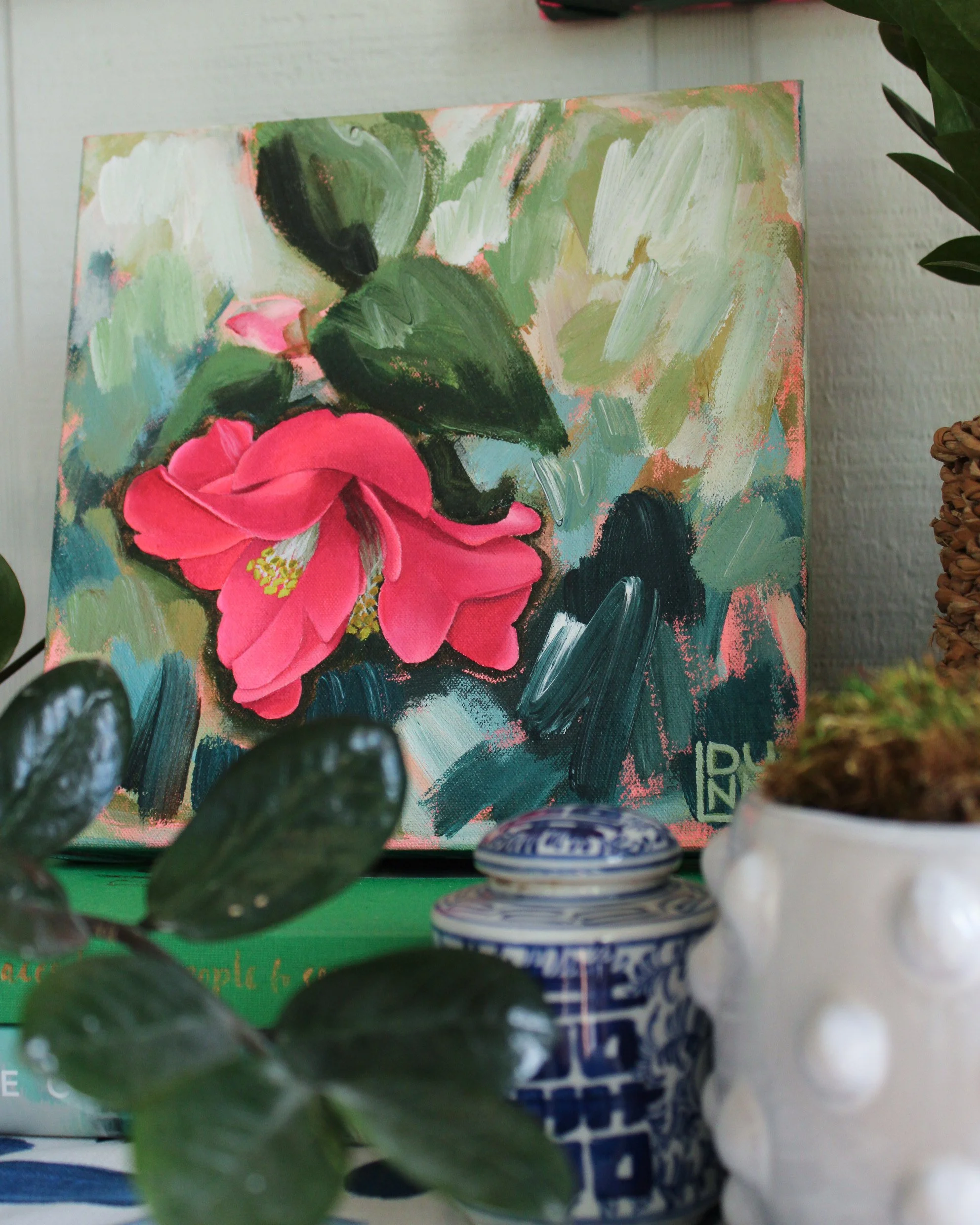 A small, colorful painting of pink flowers with green leaves, surrounded by potted plants and decorative containers on a shelf.