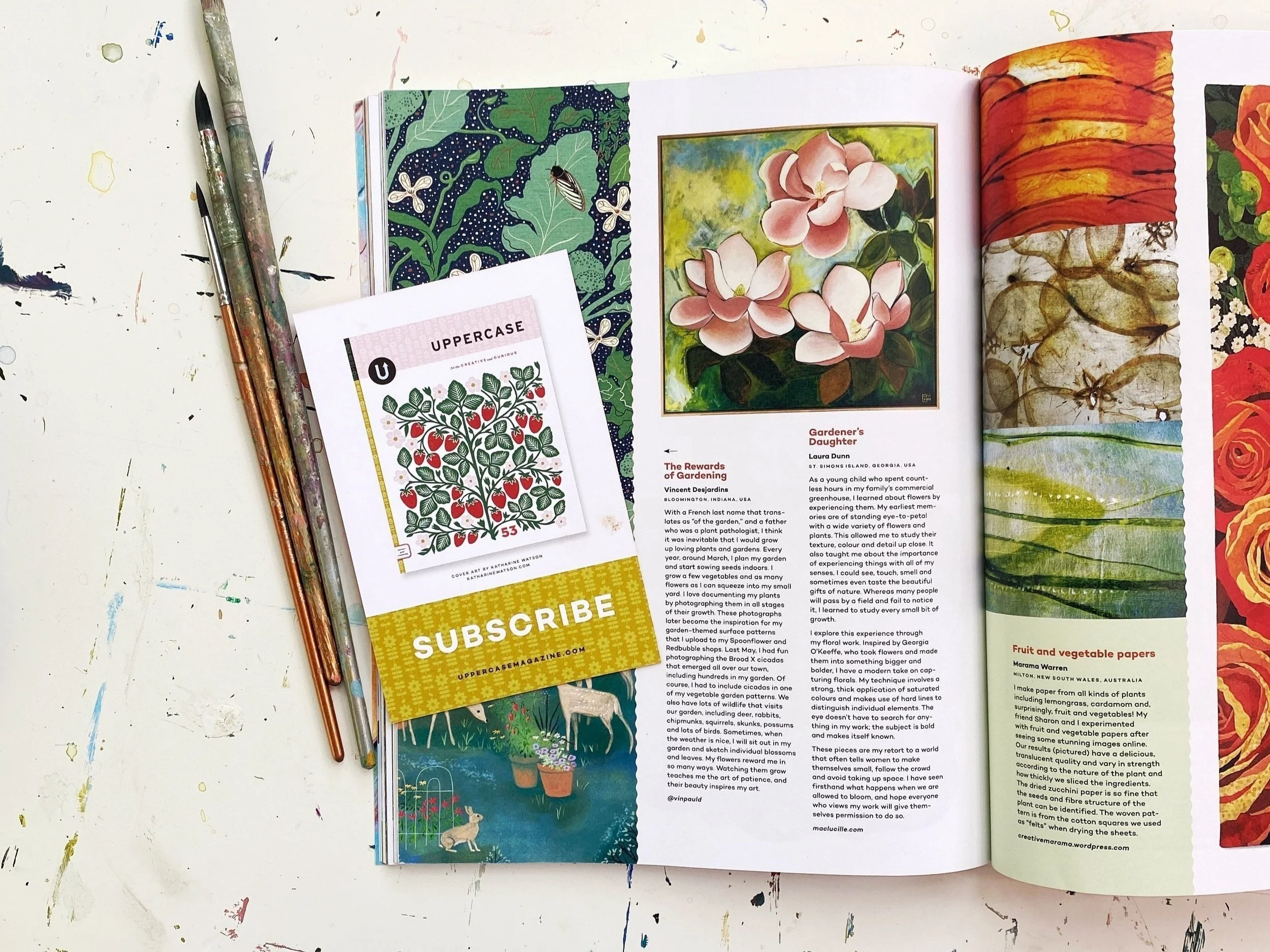 Open magazine on a paint-splattered white table, displaying botanical articles, colorful flower illustrations, an upper case alphabet chart, and gardening photos; beside it are three paintbrushes.