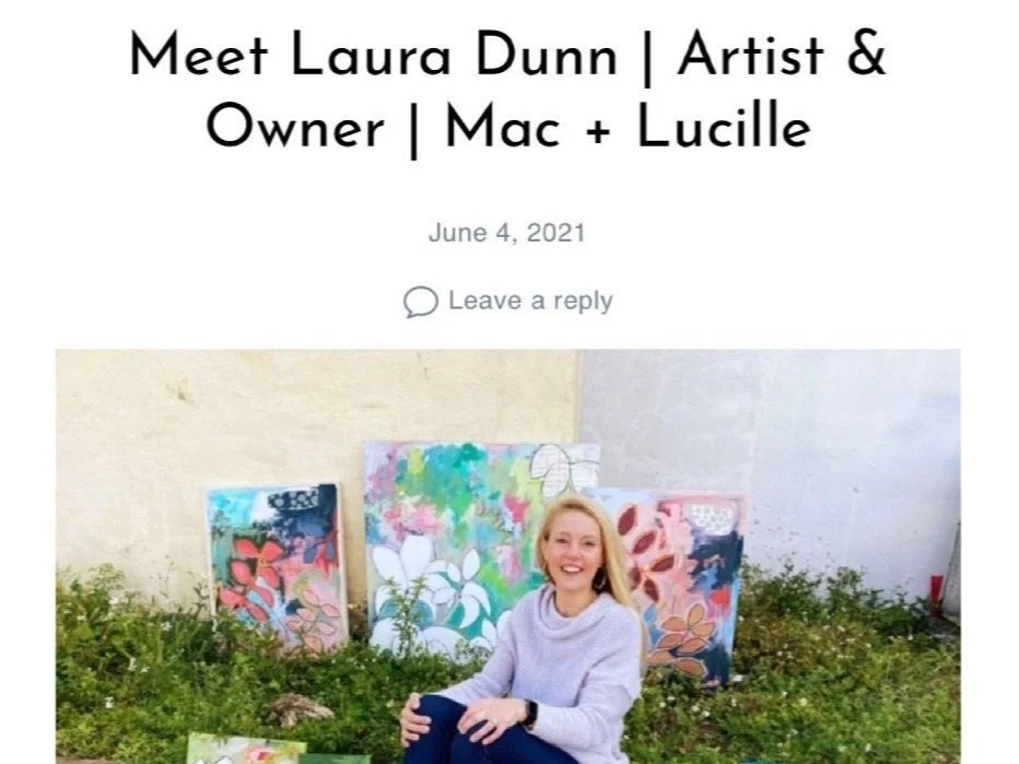 Meet Laura Dunn, artist and owner of Mac + Lucille, sitting outdoors in front of colorful abstract paintings on the ground, smiling at the camera, with greenery around her.