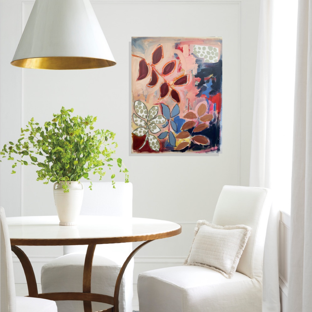 A white dining room with a round wooden table, white upholstered chairs, a green potted plant, a white wall with a colorful abstract leaf painting, and natural light coming through a window with white curtains.