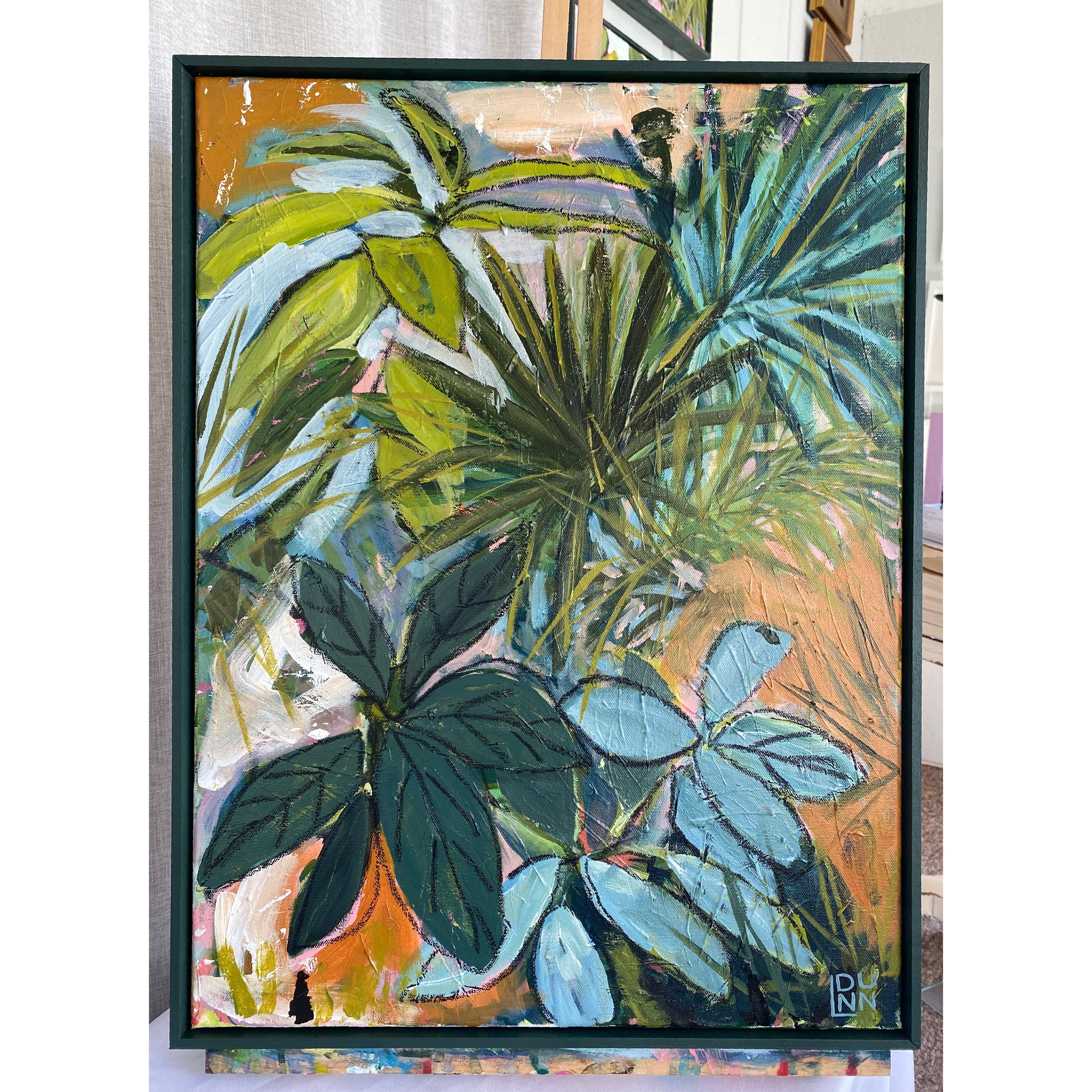 A colorful painting of lush green plants with broad leaves, framed and displayed indoors. The background features warm tones with abstract strokes.
