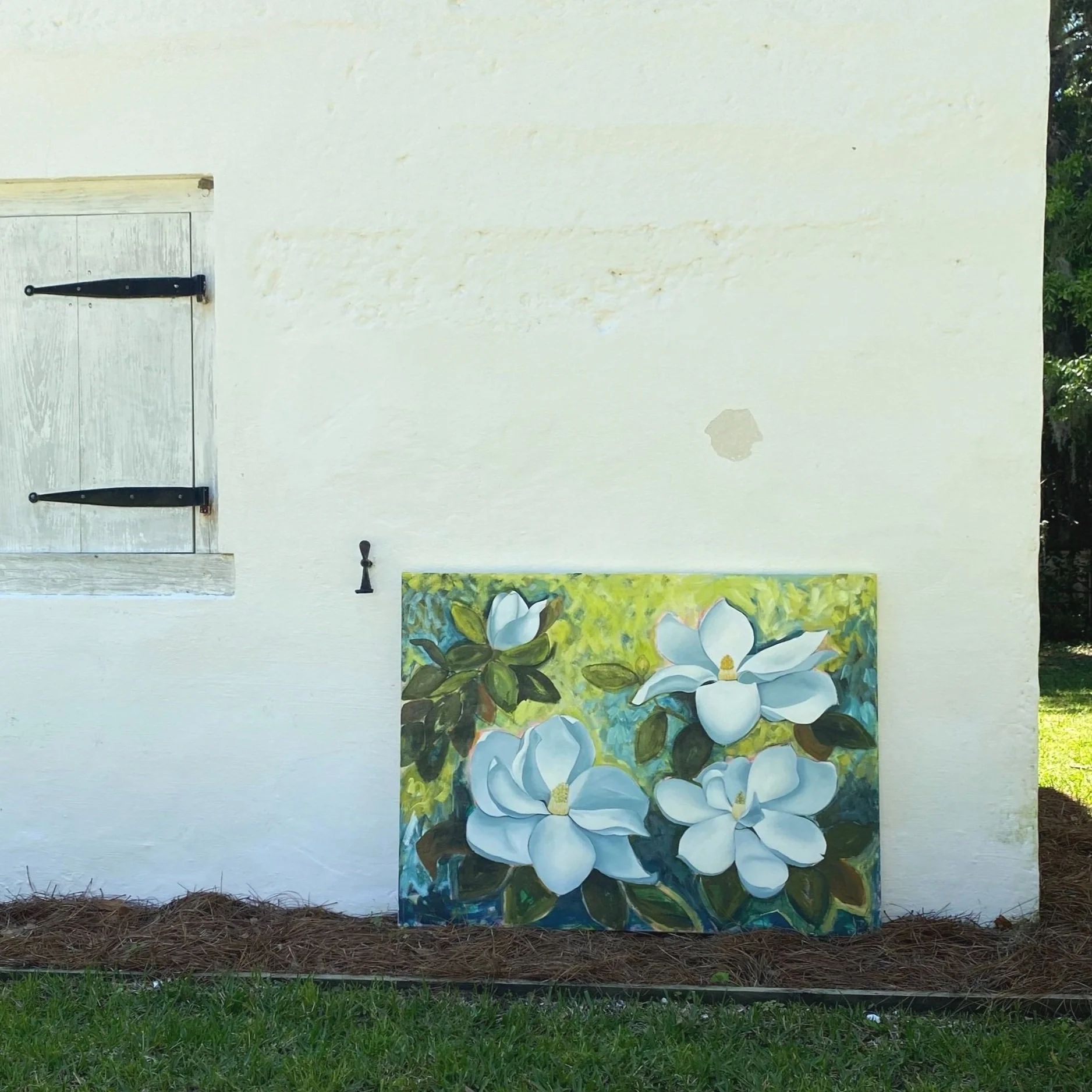 A floral painting of white magnolia flowers leaning against a white wall with a small window and hanging black hinges.