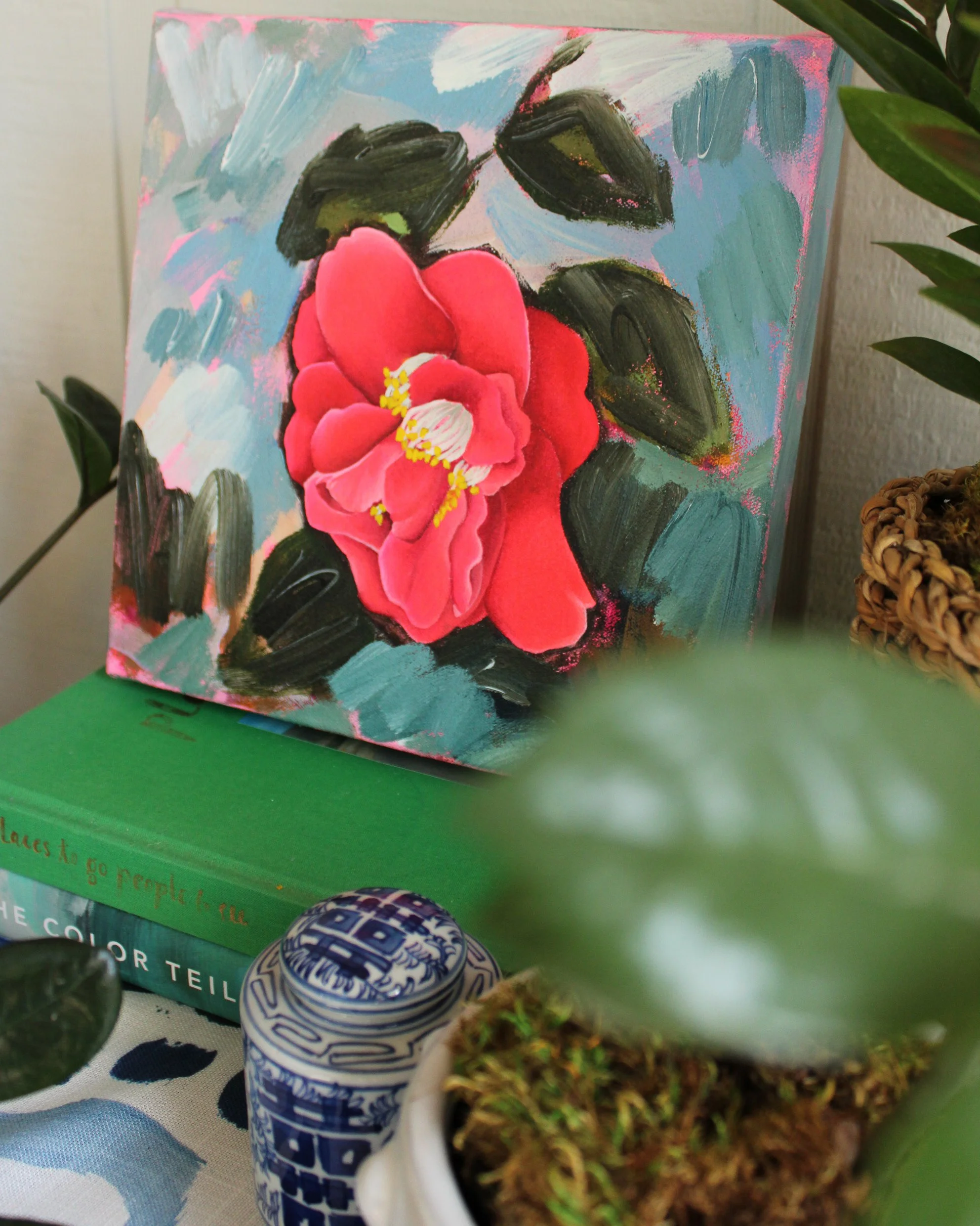 A small painted canvas featuring a pink flower with yellow stamens, black leaves, against a colorful background, placed on top of a green book. Nearby is a decorative blue and white container and some potted plants with green leaves.