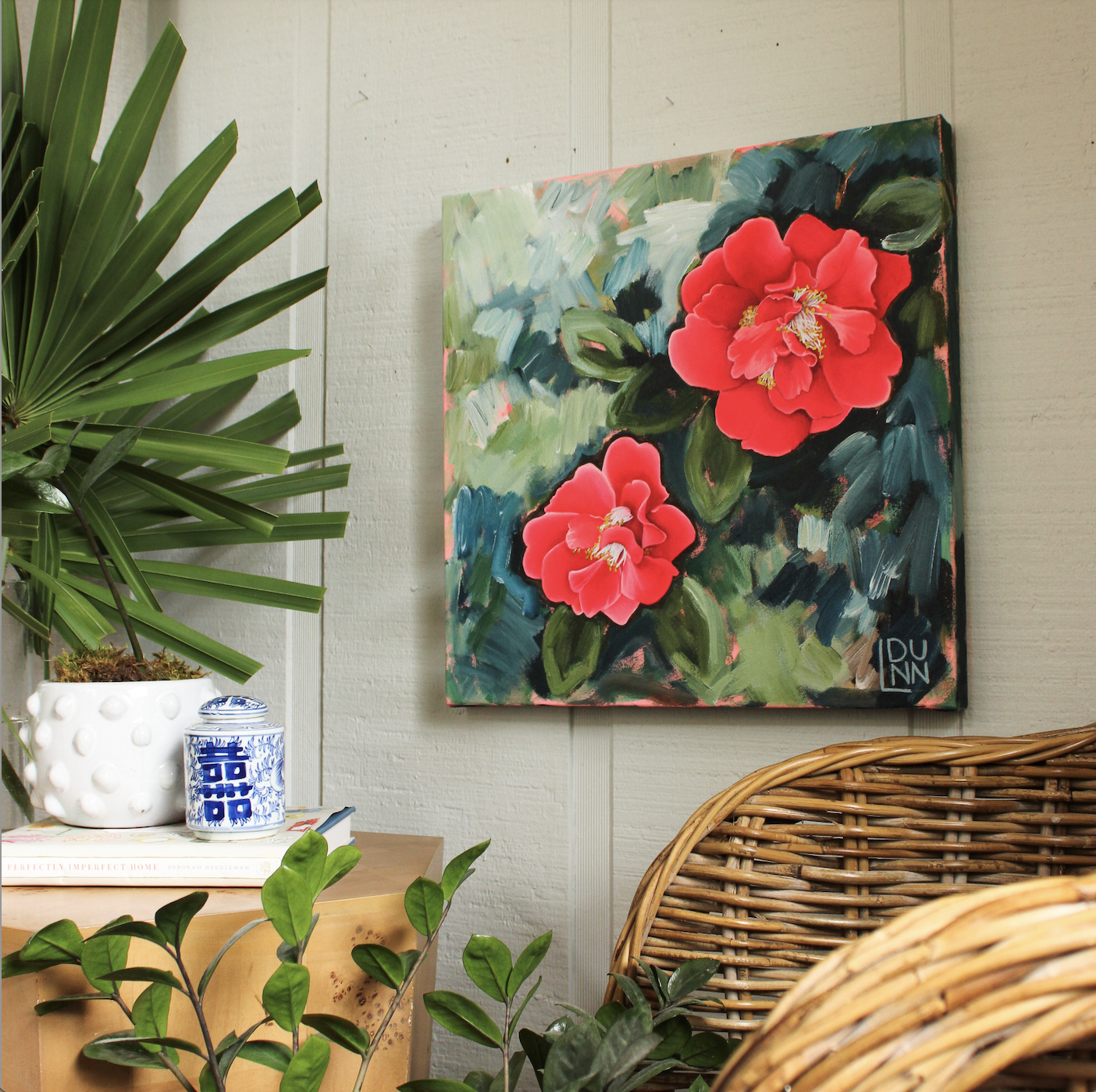 A colorful painting of red camellia flowers on a wall next to green plants and wicker furniture.