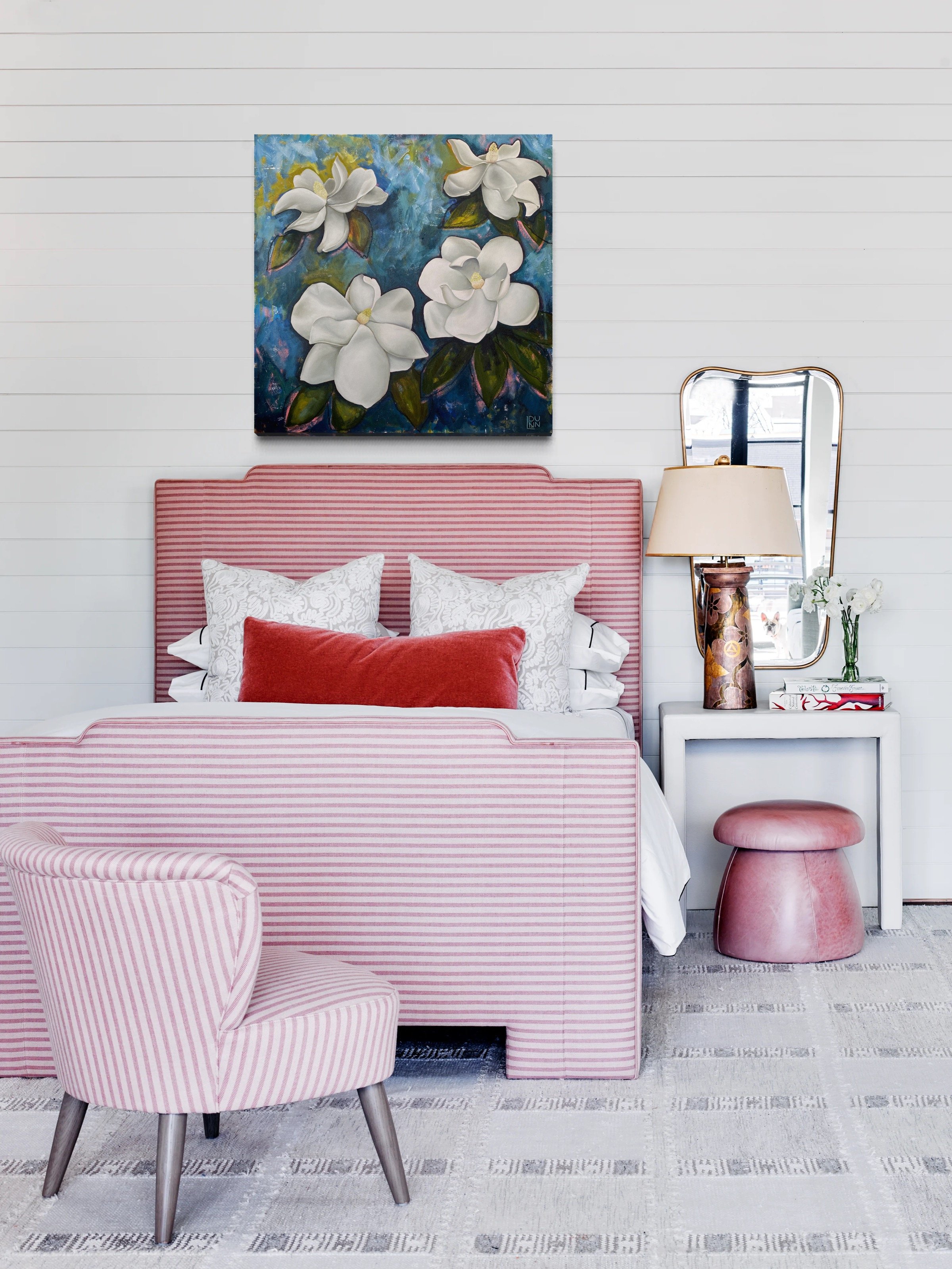 A bedroom with a pink striped bed, a white nightstand, a small pink ottoman, striped armchair, a painting of white flowers on a blue background, a large mirror, and a table lamp. The room has white walls and modern decor.