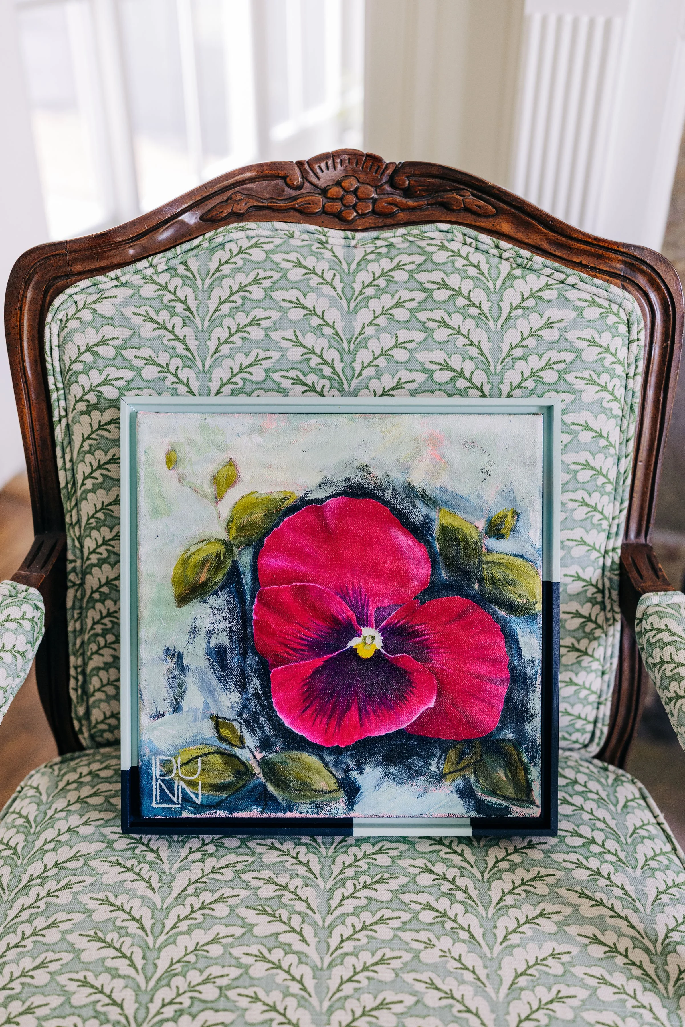 A framed painting of a large red and purple Pansy flower with green leaves resting on a green and white patterned upholstered chair by Laura Dunn of Mac Lucille.