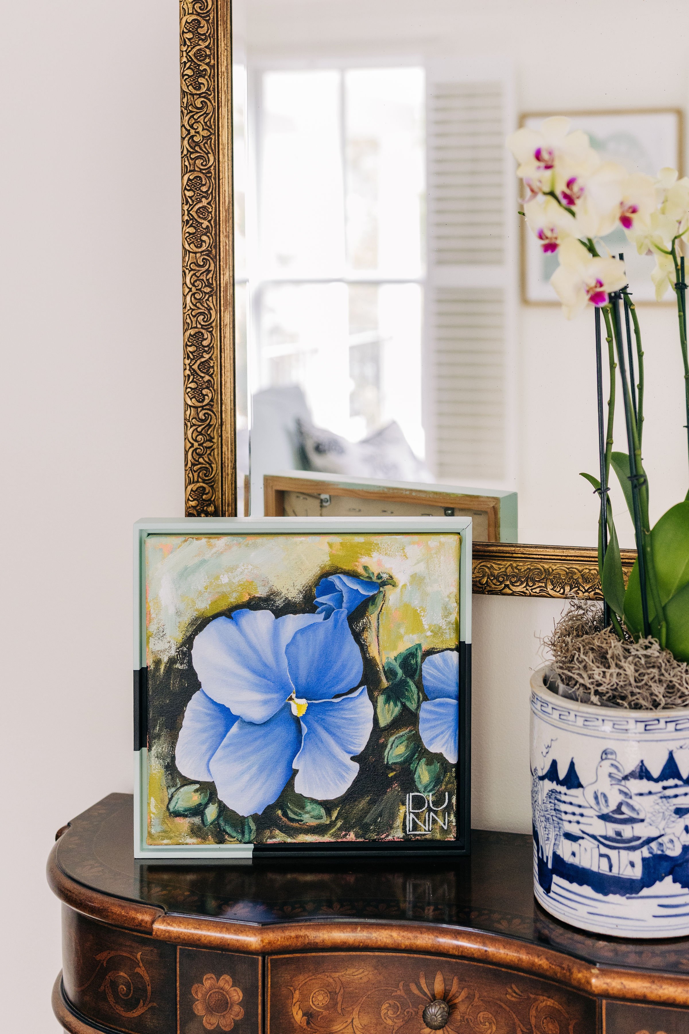 A decorative wooden table with a framed painting of blue Pansy flowers, a potted orchid plant, and a mirror on the wall reflecting a bright room with a window. Painting by Laura Dunn available with the Campbell Collective.