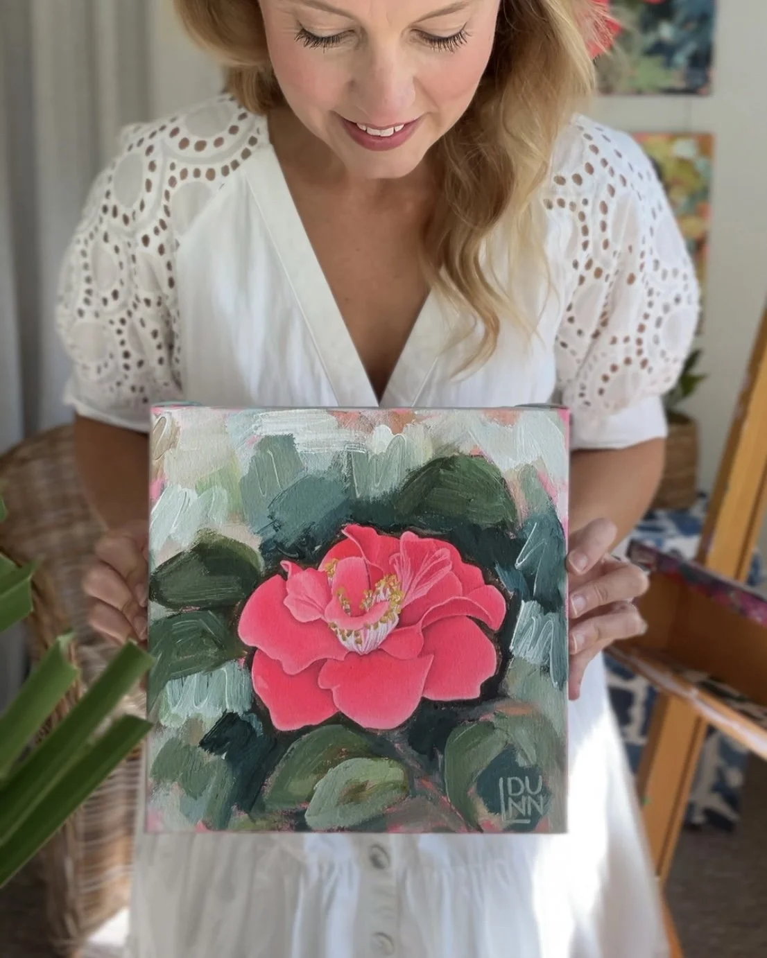Laura Dunn of Mac Lucille in a white dress holding a small painting of a pink flower with green leaves, signed in the bottom right corner.