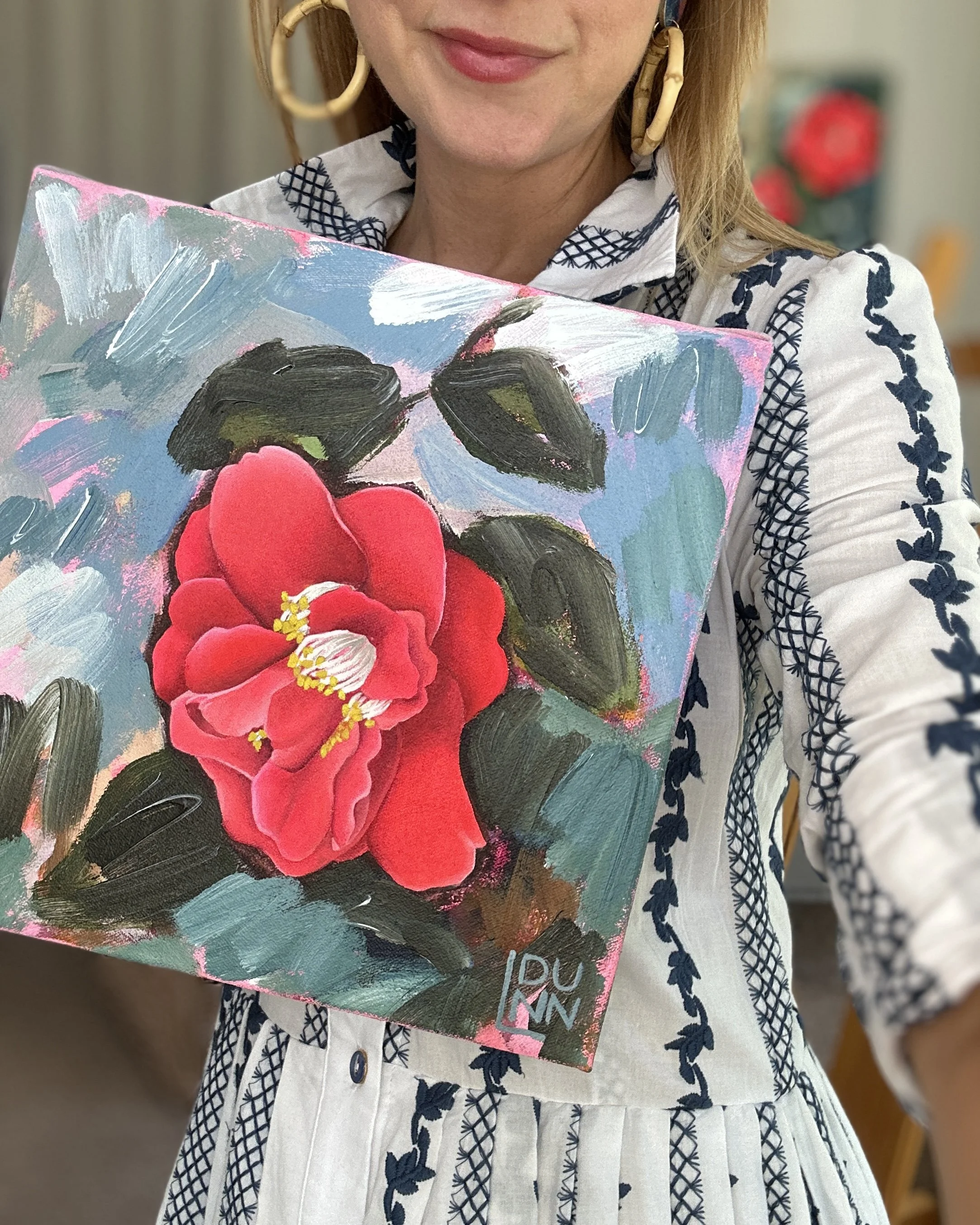 Laura Dunn of Mac Lucille holding a colorful painting of a red camellia flower with dark green leaves, against a light blue and pink background.