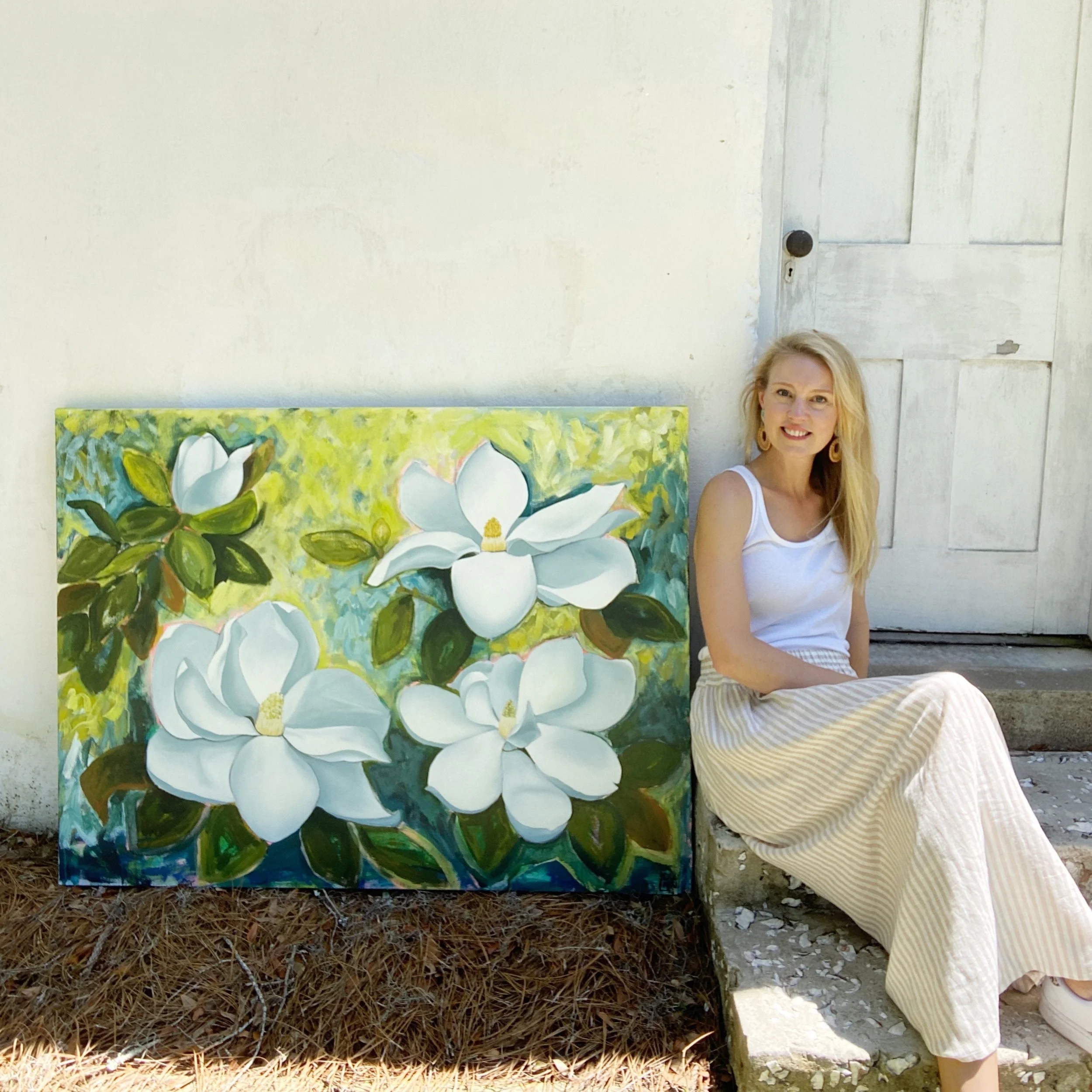 Laura Dunn sitting outside beside a completed commission painting of Magnolia blooms with green leaves on a light-colored wall with a wooden door.