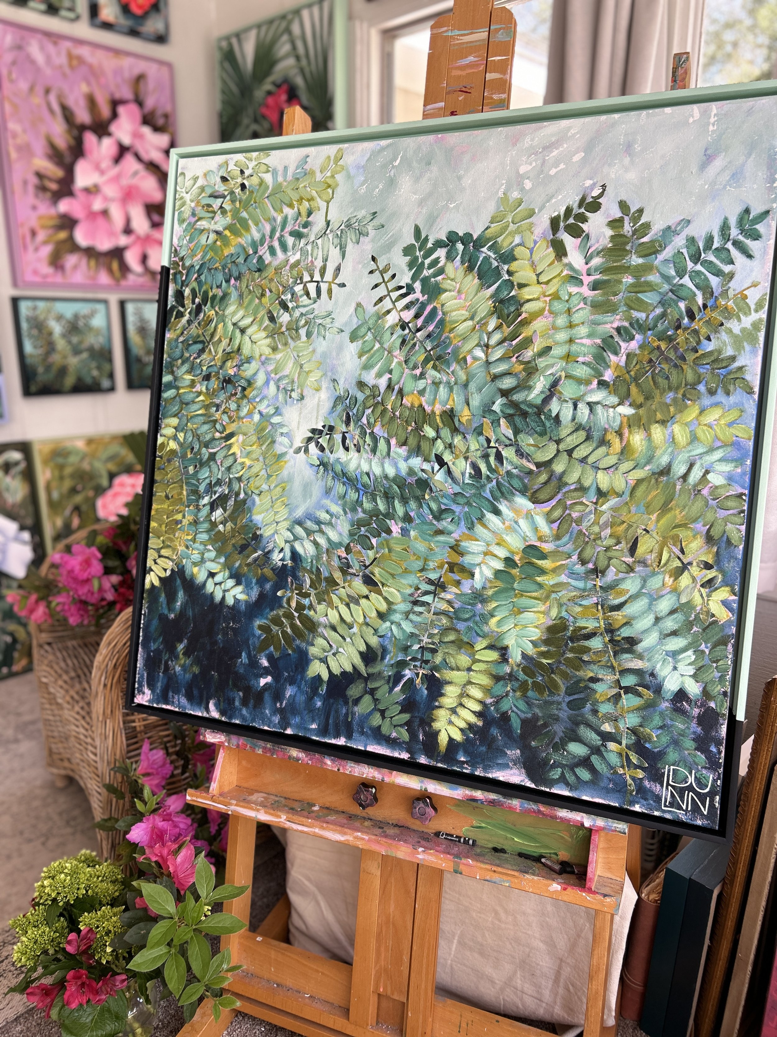 Painting of green fern leaves on a canvas, displayed on an easel in an art gallery or studio with pink and green floral artworks and plants in the background.