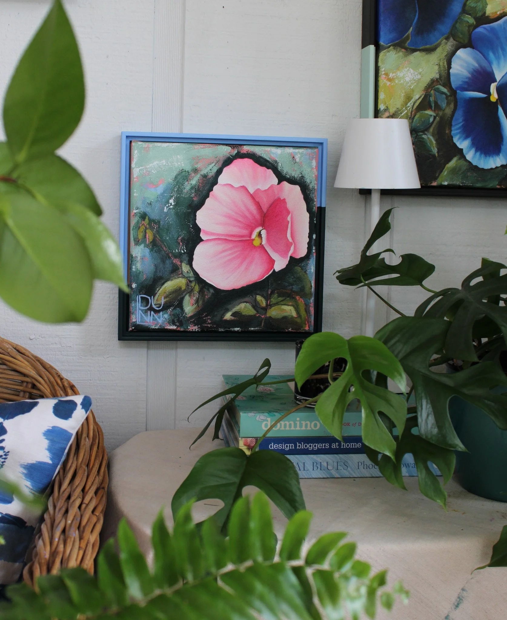 A blush-pink pansy with cream petal edges on a dark green and blue background, displayed in a blue frame among houseplants.