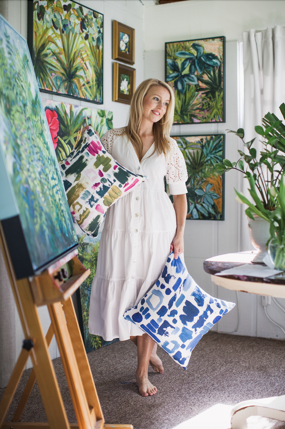 Laura Dunn of Mac Lucille in a white dress holding a colorful cushion, standing in an art studio decorated with various vibrant landscape paintings and lush green plants.