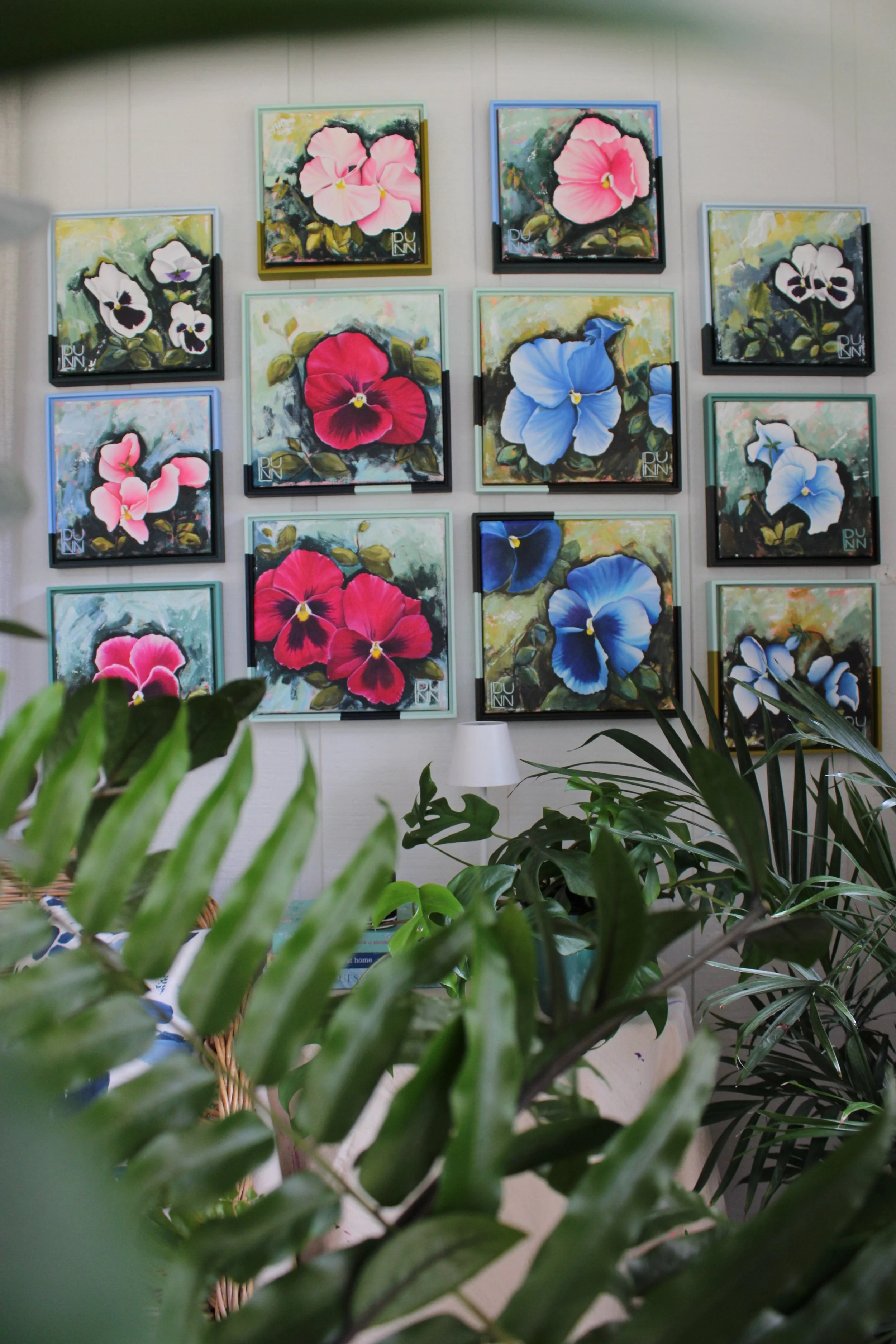A wall decorated with twelve colorful floral paintings of pansies arranged in a grid, with green houseplants in the foreground. Shown in the studio of Laura Dunn of Mac Lucille.