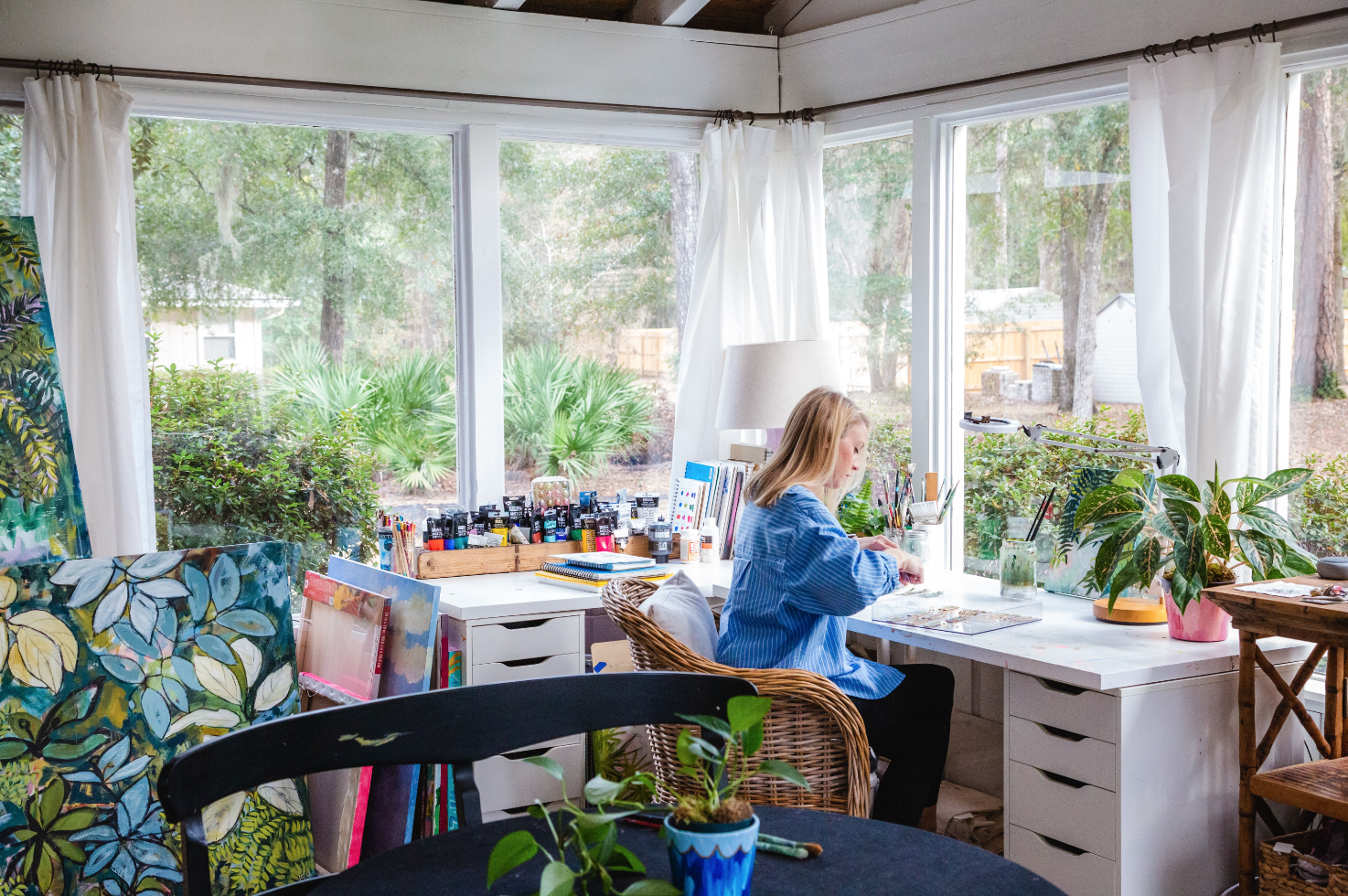 Laura Dunn sitting at a white desk in a sunroom, working on art projects with paints, brushes, and art supplies, surrounded by plants, artwork, and large windows revealing a green backyard.