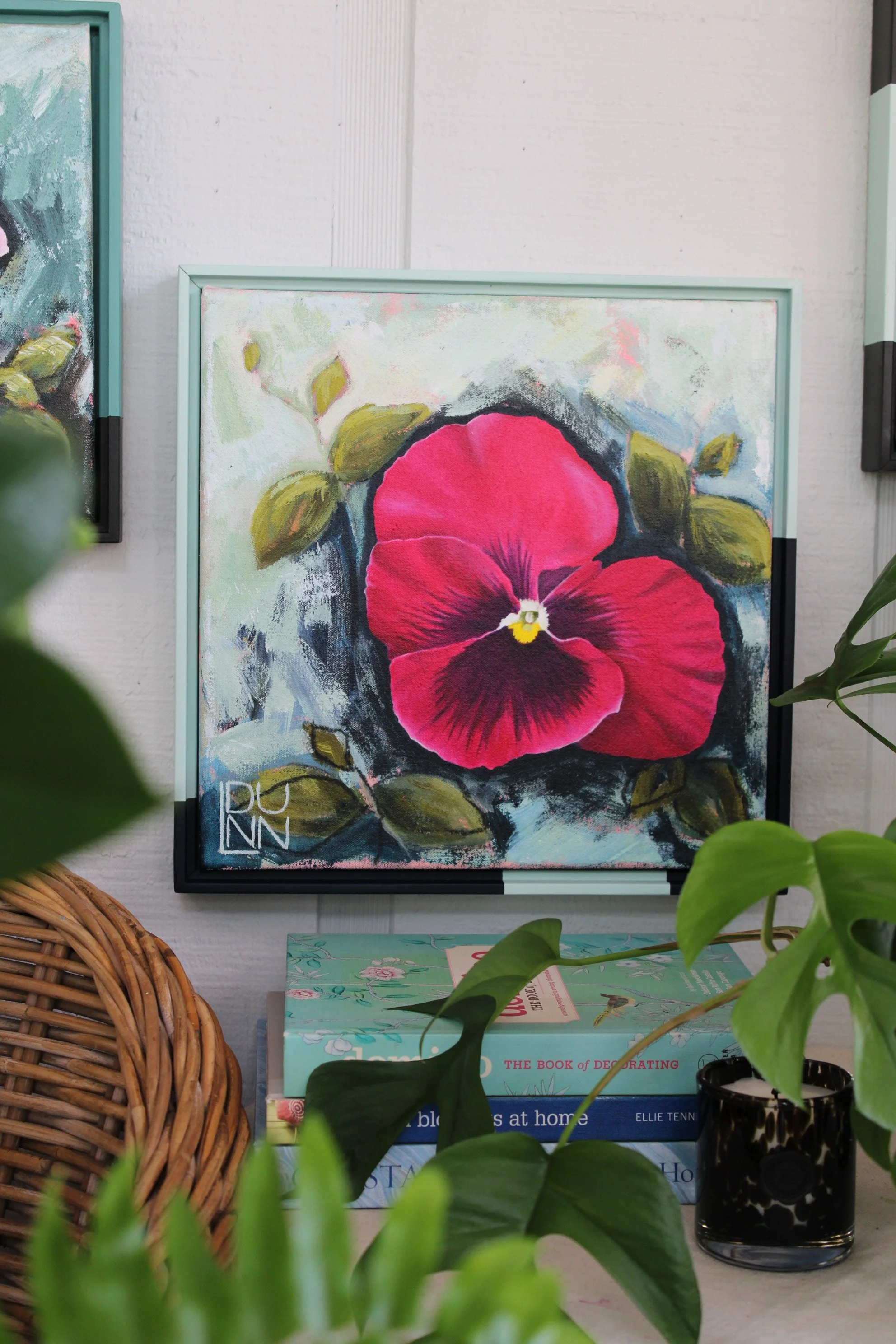 A single raspberry-pink pansy with dark burgundy markings on a pale green and blue background, framed above a stack of books.