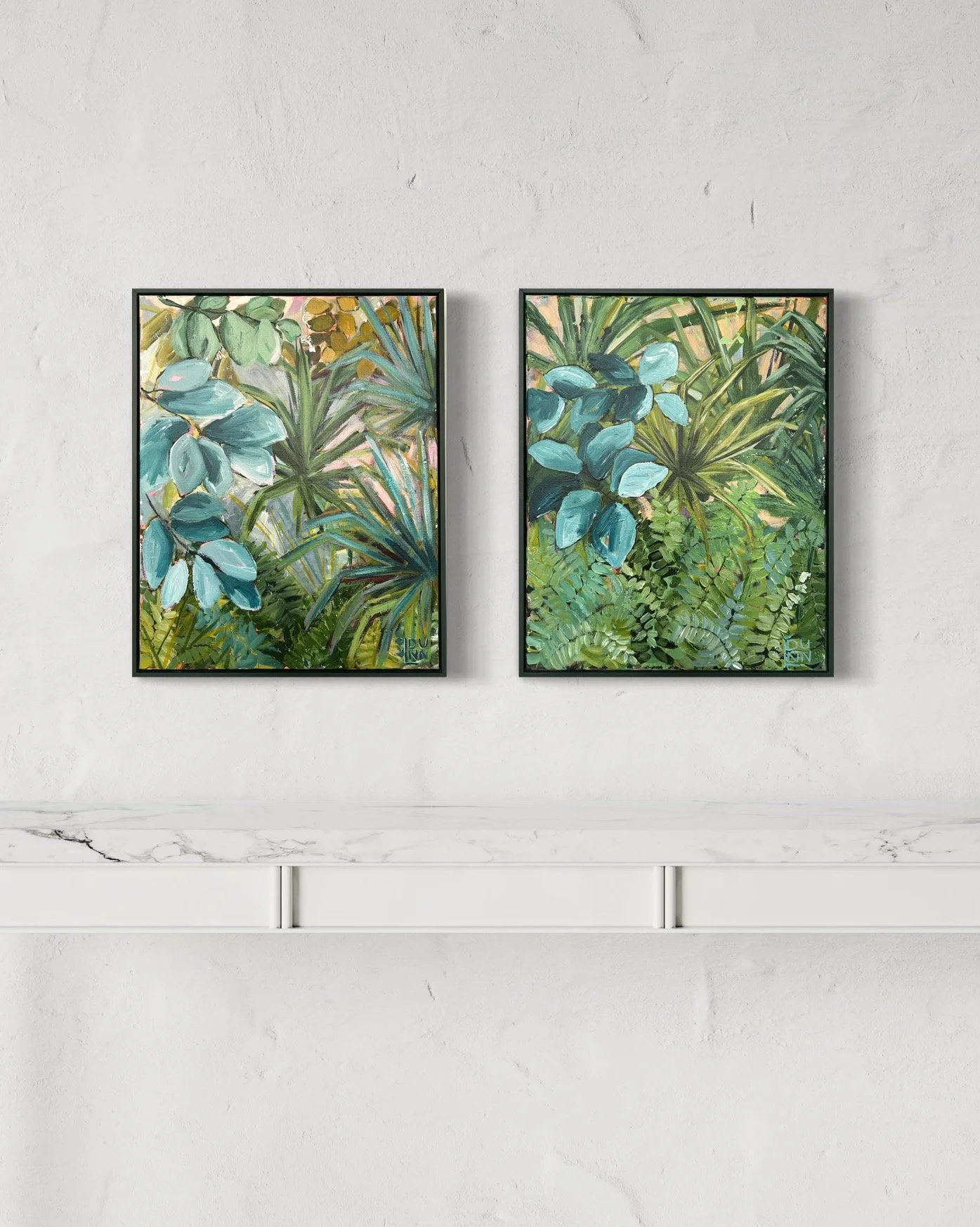Two framed paintings of lush green and blue tropical plants hanging on a white wall above a white marble shelf.