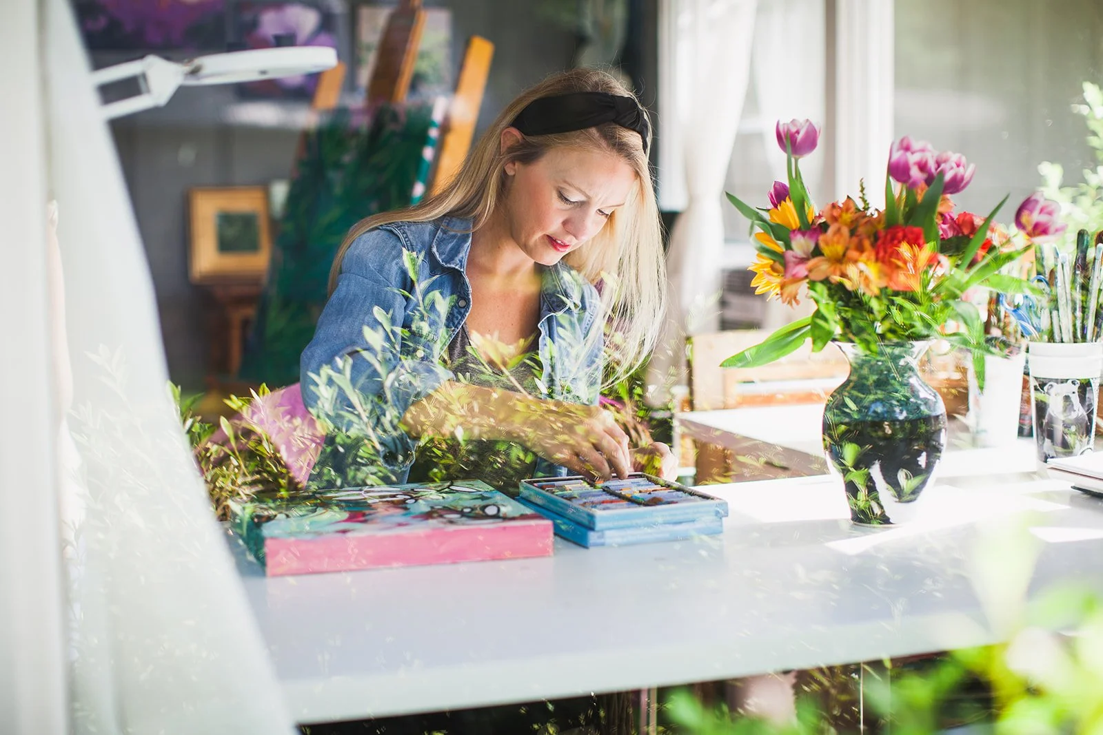 Laura Dunn of Mac Lucille working on a painting project at a bright table with a large vase of colorful flowers and art supplies.