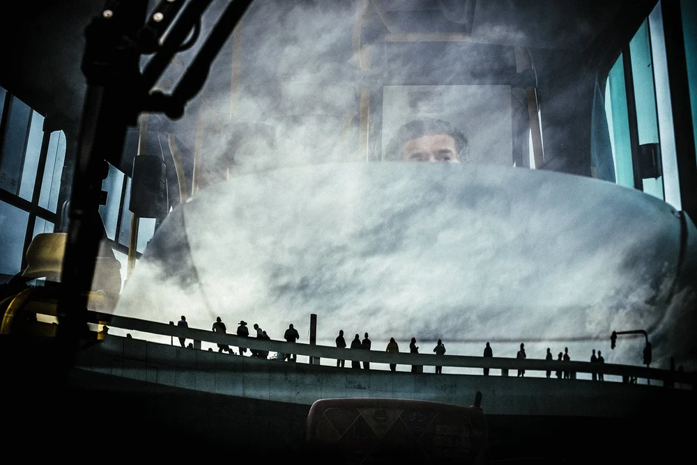 A bus driver's face, lit and half-swallowed by reflection, seen through a windscreen that mirrors an enormous cumulus cloud filling the whole glass; a row of tiny black commuter silhouettes walks a raised rail along the bottom third