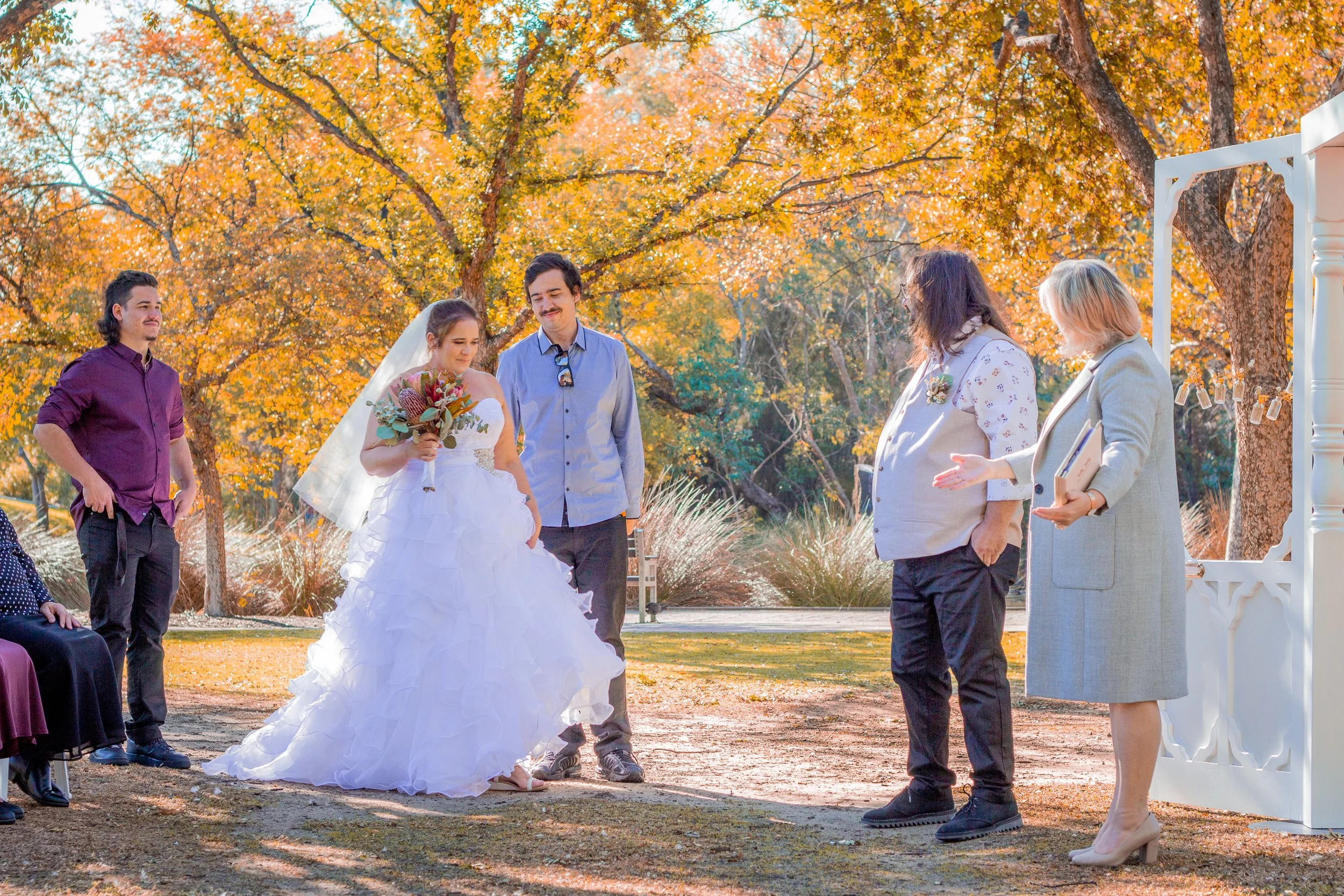 A couple's outdoor wedding ceremony with a bride in a white wedding dress holding a bouquet, standing beside a groom in a light blue shirt. Two guests, one with long hair and a gray blazer, the other with curly hair and glasses, stand to the right. A man in a purple shirt and a woman in a dark dress are visible on the left. The background features orange autumn leaves and trees.