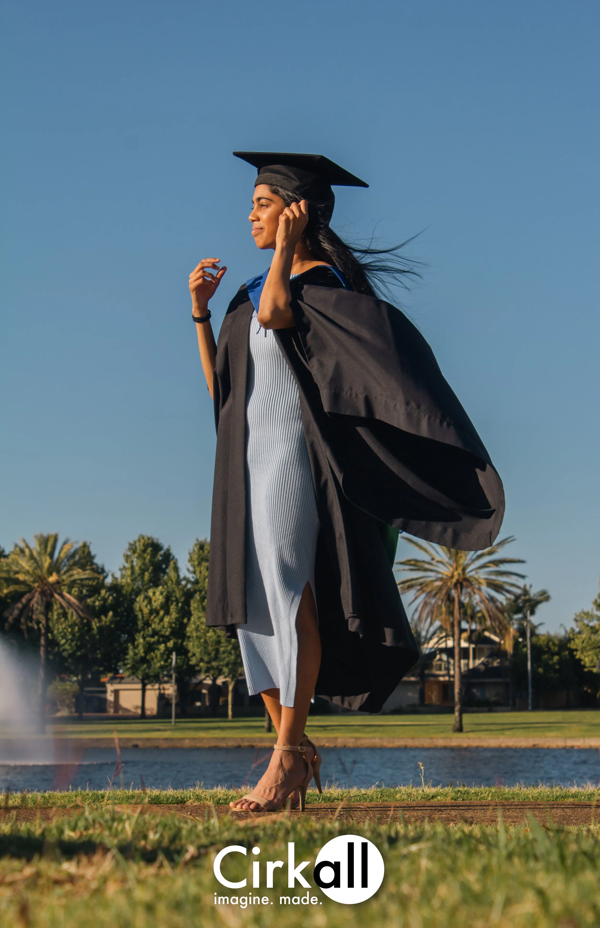 A young woman in a graduation cap and gown standing outdoors near a lake with palm trees, celebrating graduation.