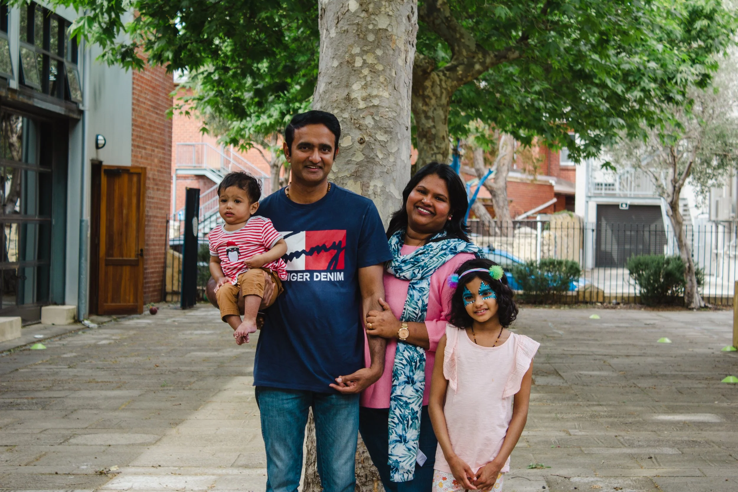 A family of four standing outdoors under a tree, smiling at the camera. The father is holding a toddler, the mother is smiling with a child in front of her. All are casually dressed.