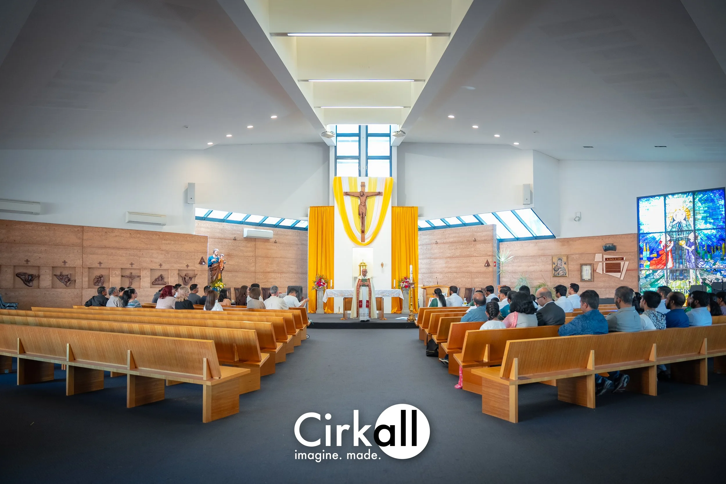 Interior of a church with congregation seated on wooden pews, altar with yellow drapes, crucifix, and stained glass window.