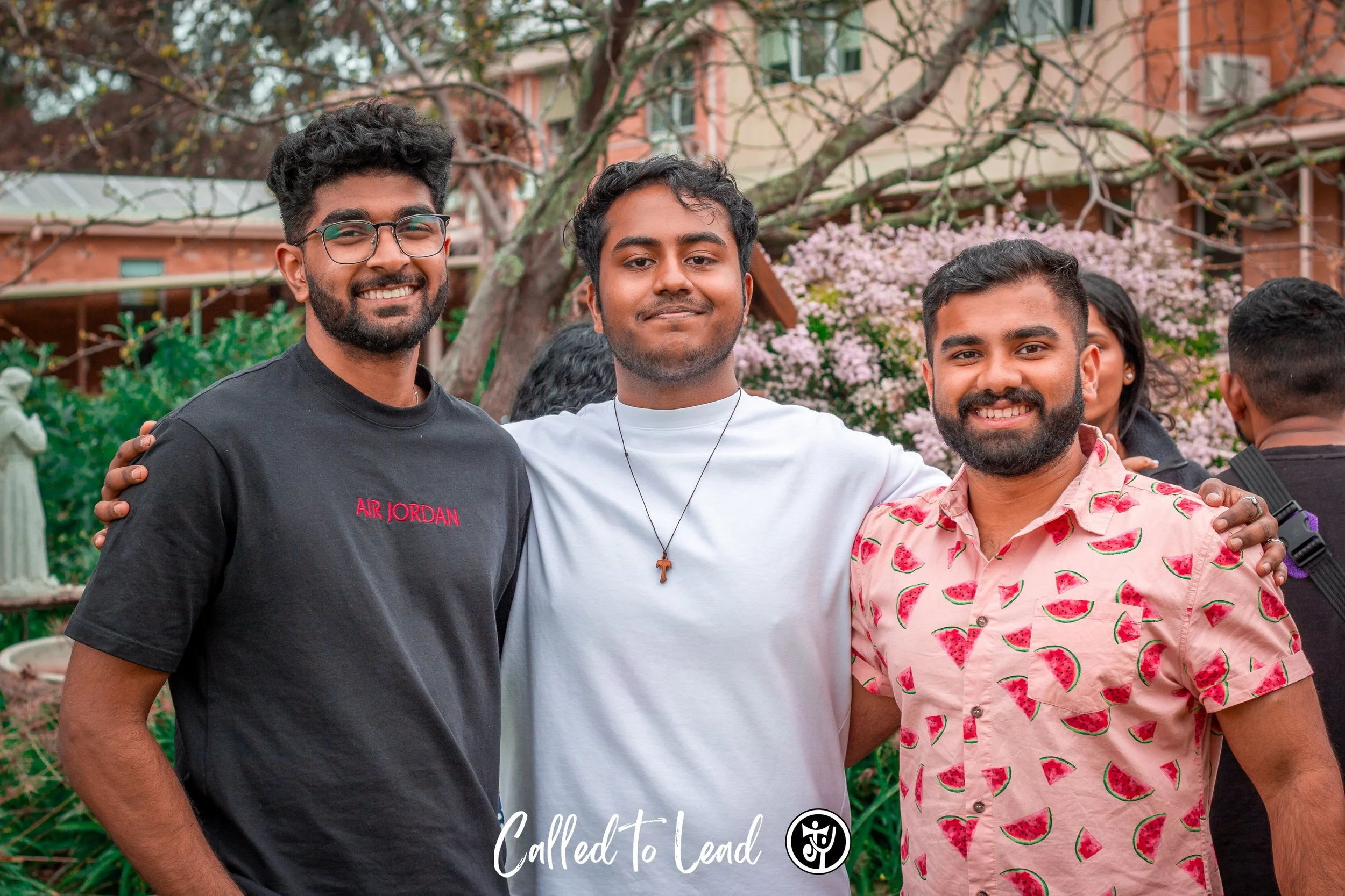 Three smiling young men standing together outdoors with their arms around each other. Behind them is a flowering tree and a building, suggesting a park or garden setting.