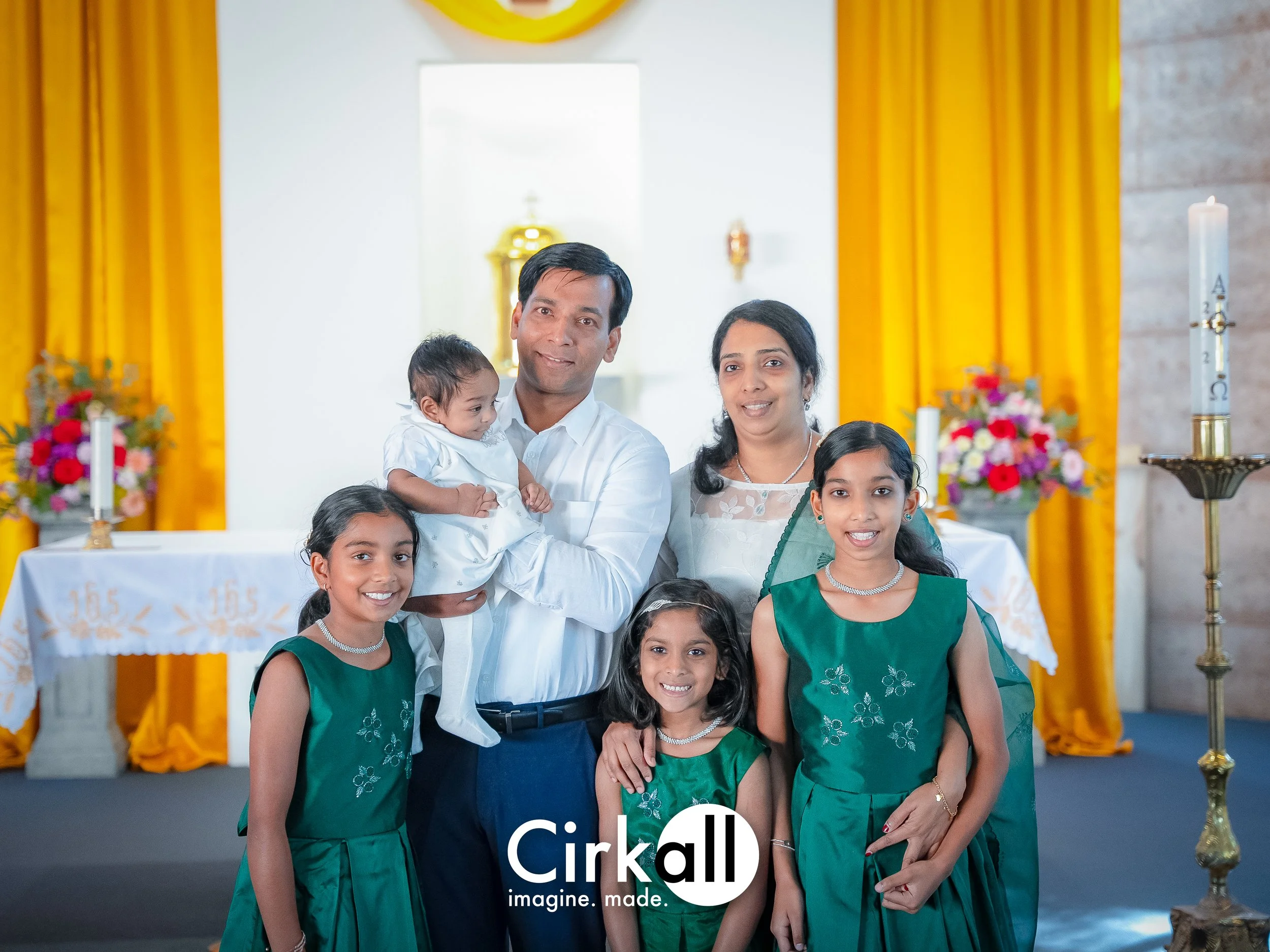 A family of seven, including two adults and five children, posed together inside a church decorated with yellow curtains, flowers, and candles. They are dressed in formal attire, with the children wearing green dresses.